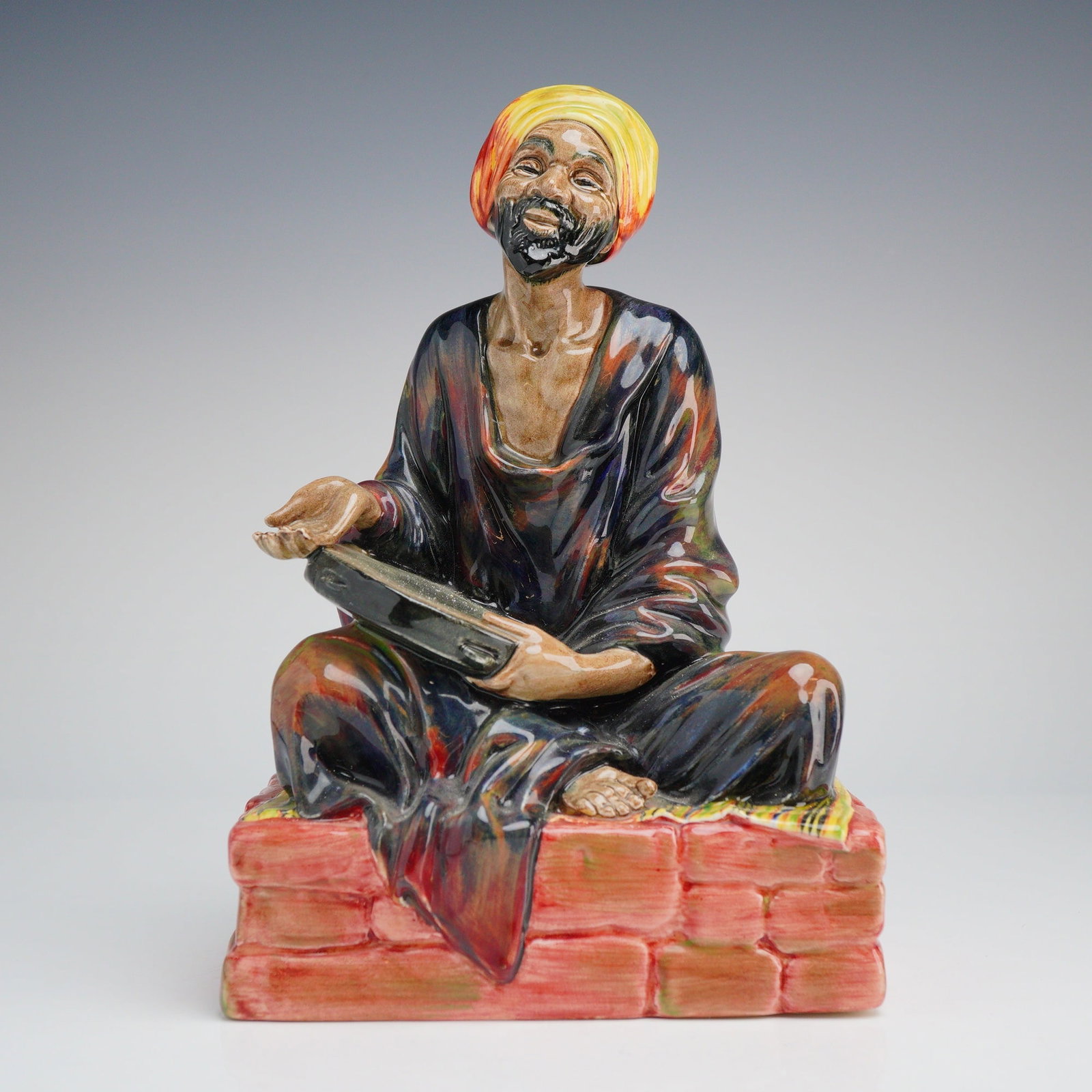 Royal Doulton Porcelain Figurine, Mendicant HN1365: A Royal Doulton glazed porcelain figurine titled The Mendicant, identification number HN 1365. The figure depicts a man with a black beard and a yellow and orange turban, seated cross-legged on a red 