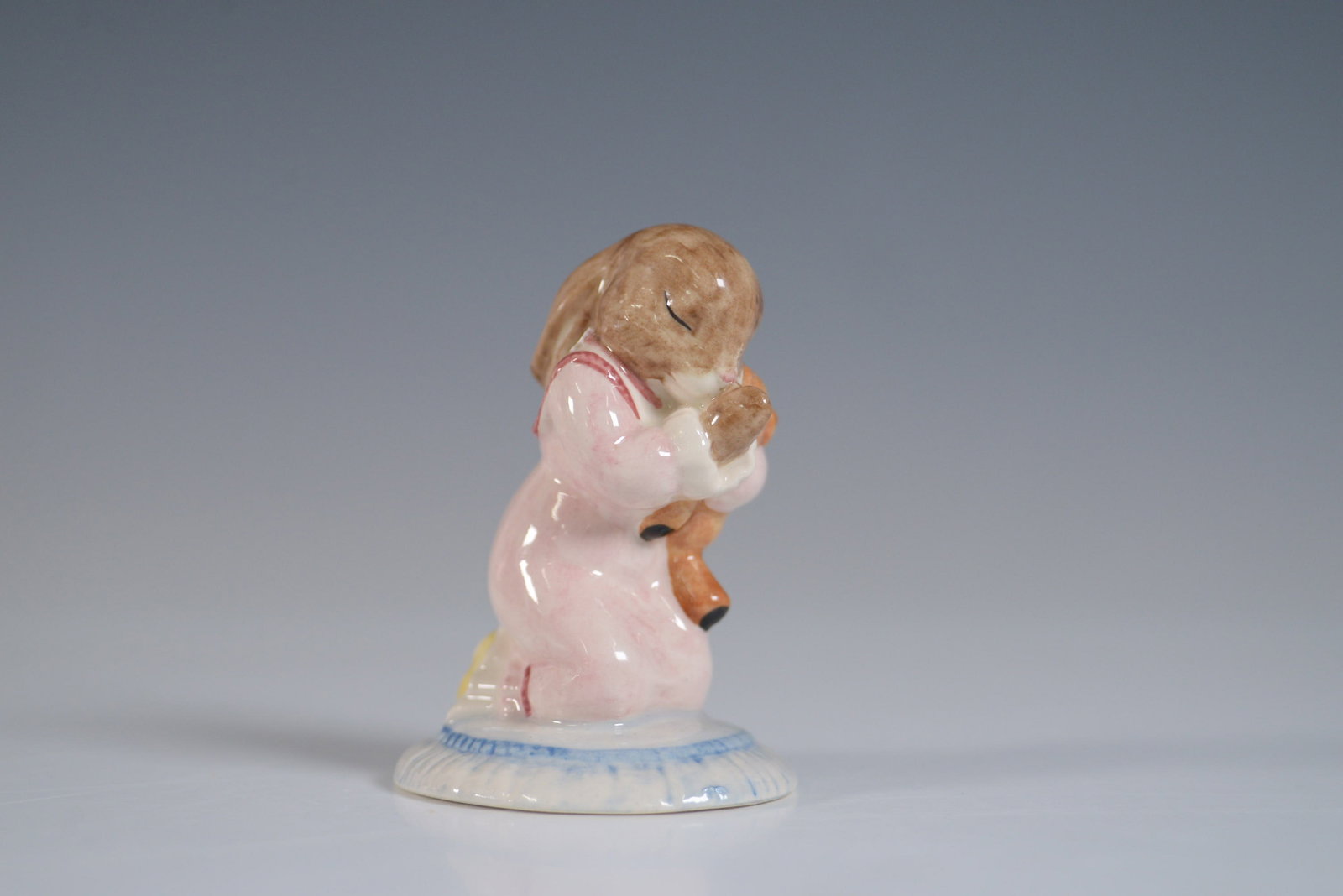 Royal Doulton Bunnykins Figurine, Goodnight, DB157: This delightful Royal Doulton Bunnykins figurine, titled Goodnight (DB157), depicts a bunny in a pink nightgown, gently cradling a teddy bear as it prepares for sleep. The figurine stands at 3"H and i