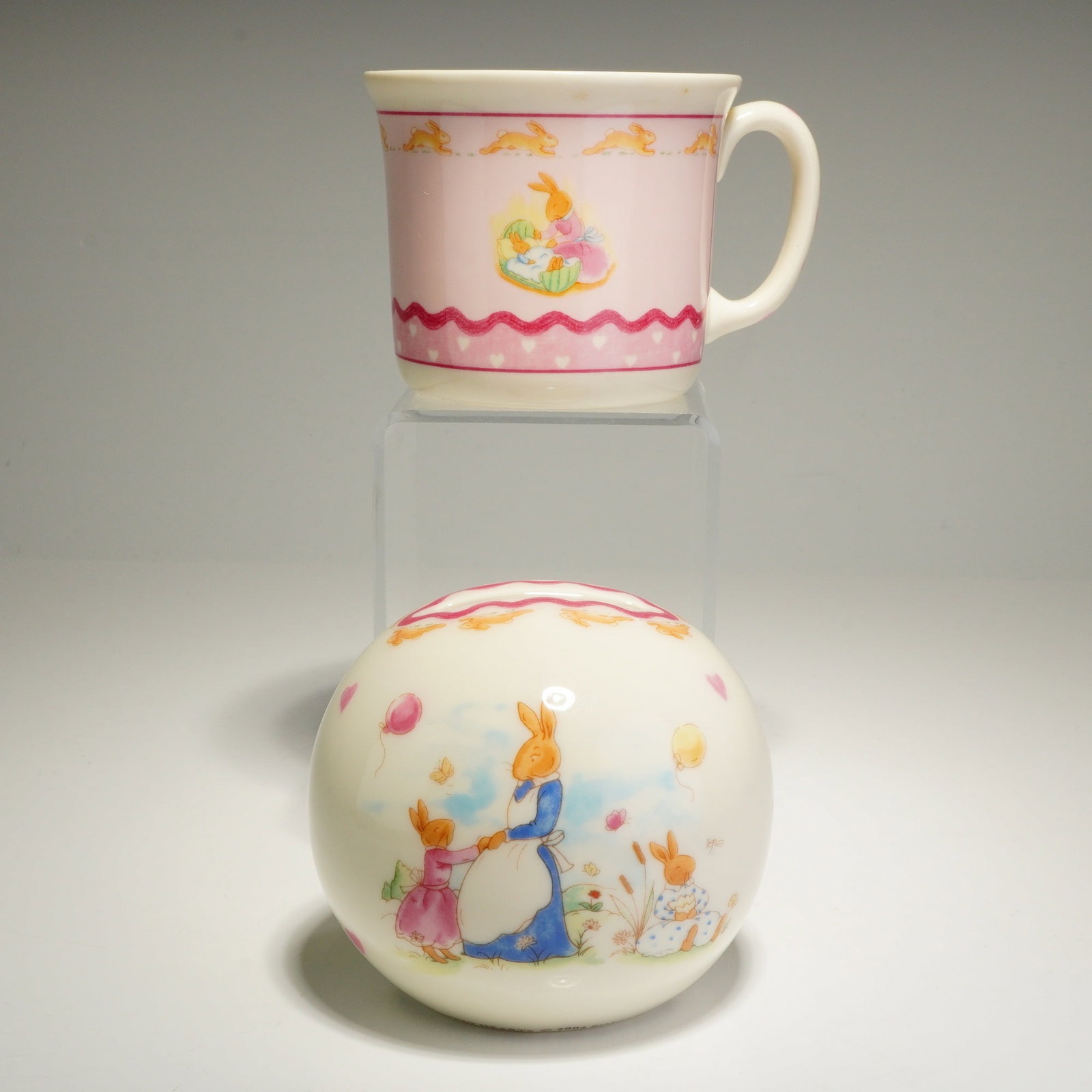 2pc Royal Doulton Bunnykins Sweet Hearts Mug and Money Bank: The handled mug and spherical money box are crafted from bone china with a cream-colored ground, while the mug features a prominent pink area as its main background. Both pieces showcase colorful illu