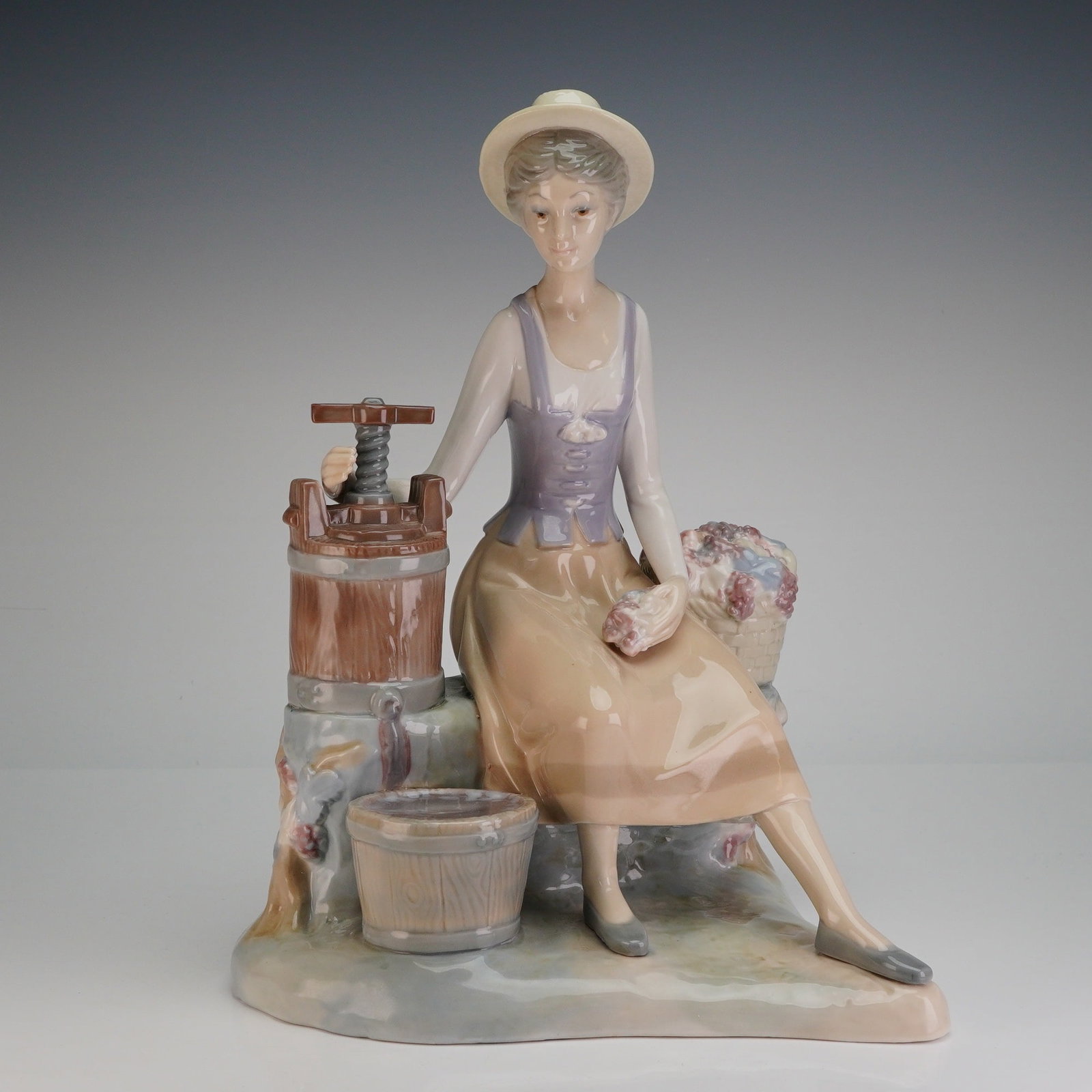 Zaphir Porcelain Sculpture, Woman with Wine Press: The piece depicts a young woman, or country lass, seated in pastoral attire next to a brown wooden fruit press. She is dressed in a wide-brimmed hat, purple bodice, and tan skirt. Beside the press is 