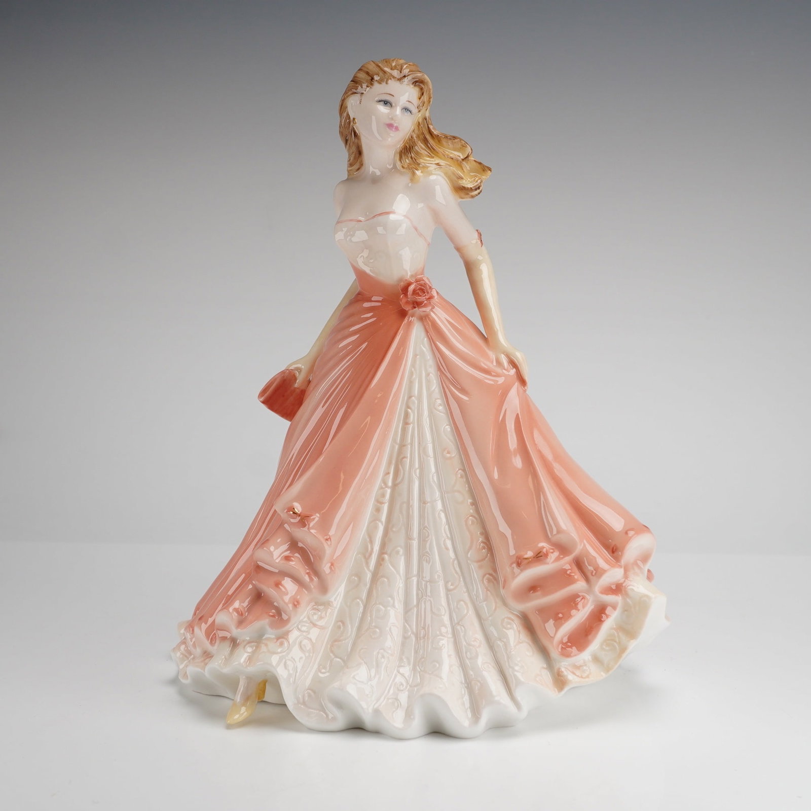 Coalport Bone China Figurine Congratulations by Jack Glynn: A Coalport bone china figurine titled Congratulations, modeled by Jack Glynn. This hand-decorated piece depicts a young woman with wind-swept blonde hair in a flowing, floor-length ballgown. The figur