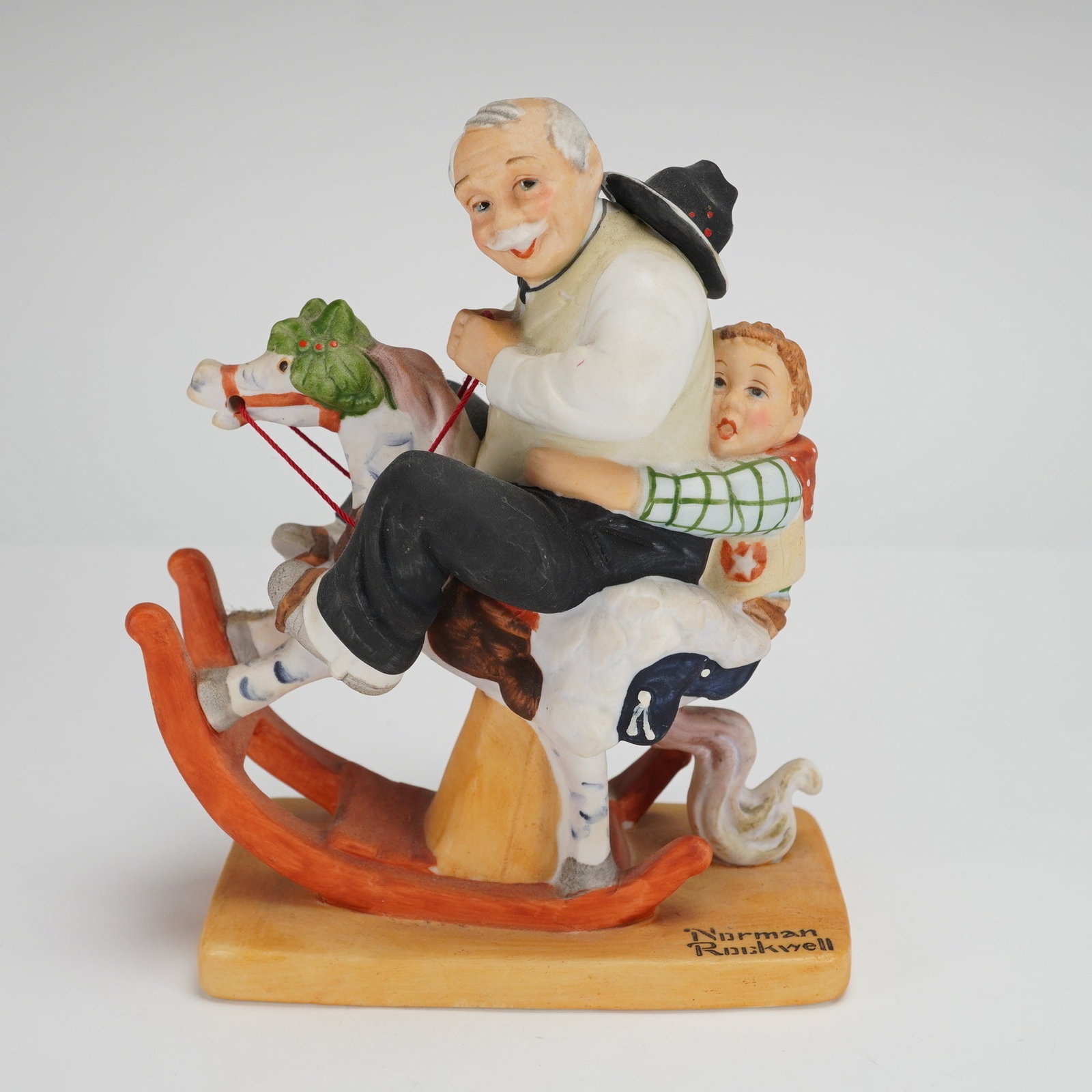 The Danbury Mint Norman Rockwell Porcelain Figurine Gramps at the Reins: This hand-painted porcelain figurine, titled Gramps at the Reins, depicts an elderly man and a young boy seated together on a rocking horse. The piece is crafted in the sentimental style associated wi