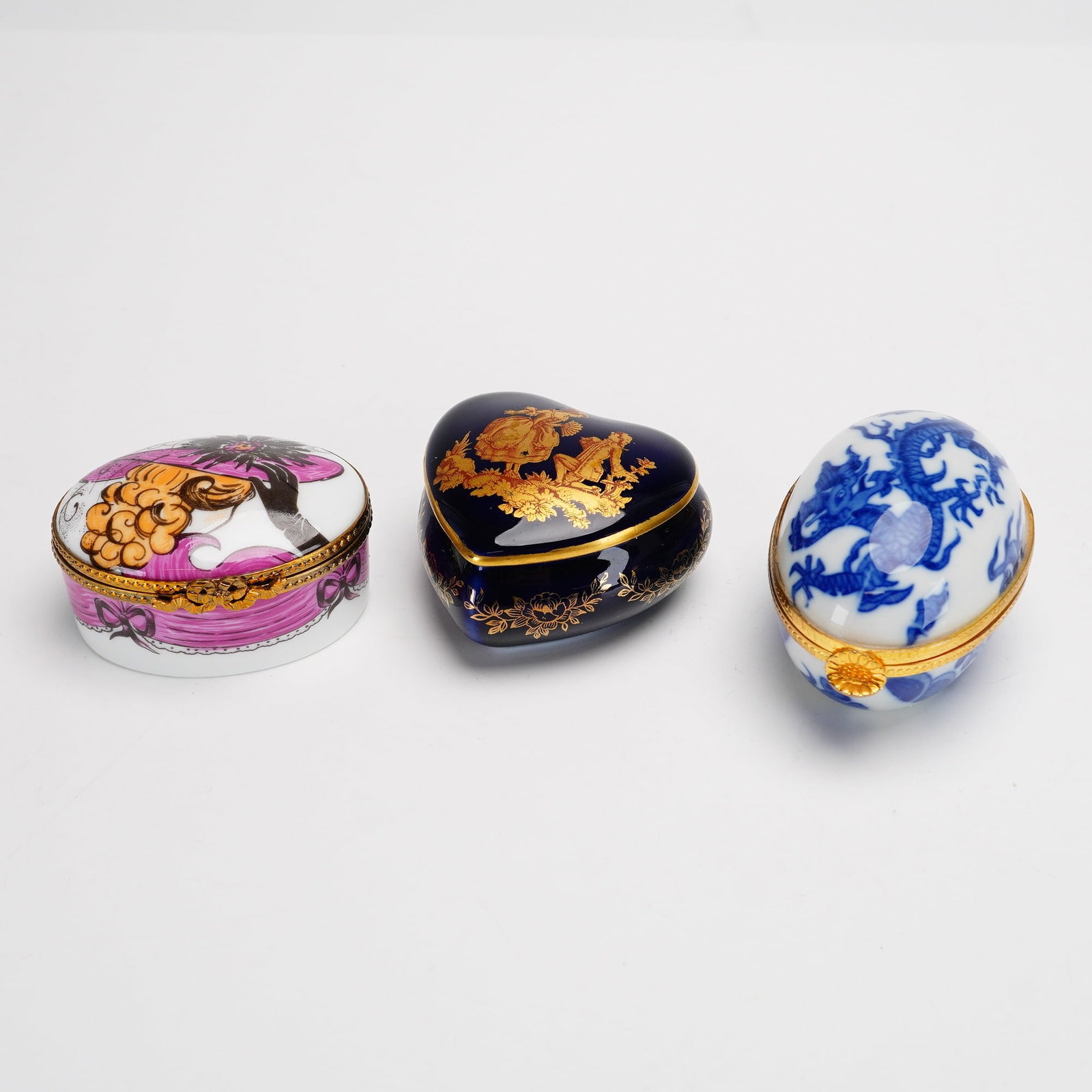 3pc Porcelain Trinket Boxes including Limoges and Neiman Marcus: This collection consists of three porcelain trinket boxes with varied decorations and forms. The first is a hinged oval box with gilt metal mounts and a floral clasp. The lid displays a transfer-print