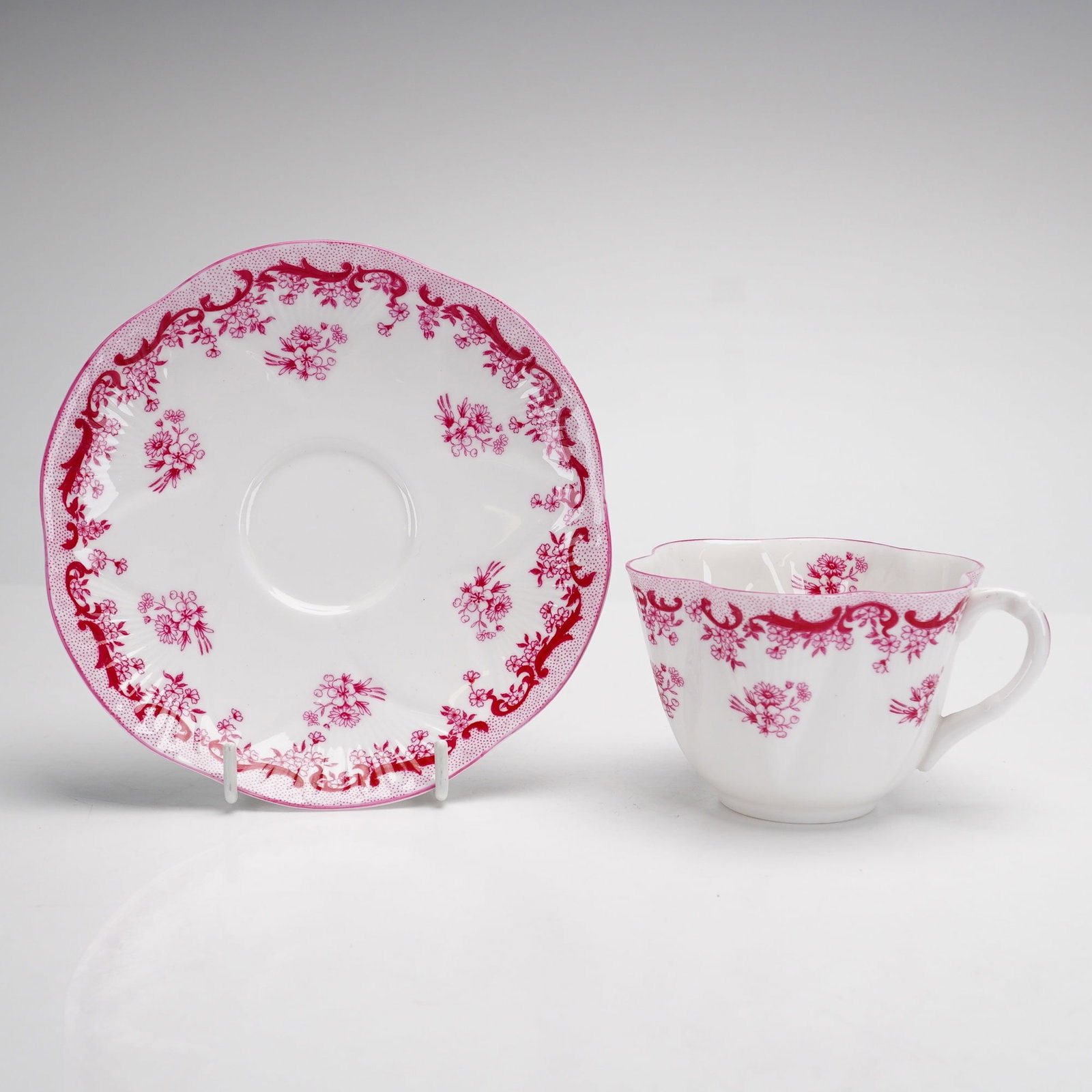 Shelley England Dainty Pink Floral Cup and Saucer Set: A two-piece fine bone china teacup and saucer set manufactured by Shelley in England. The set features the iconic Dainty shape, characterized by a fluted, lobed body and scalloped, wavy rims. The deco