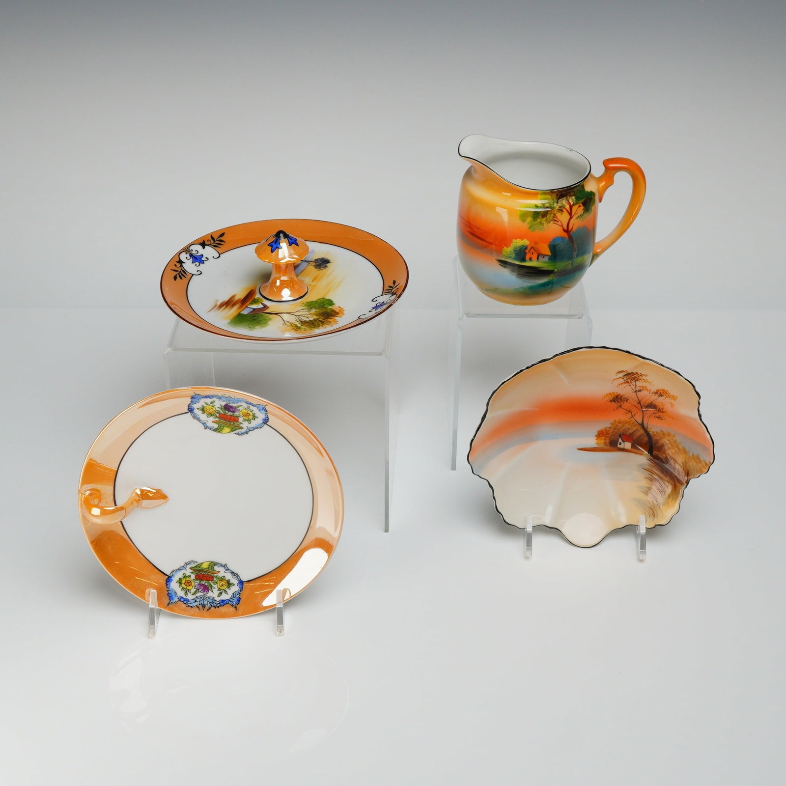 4pc Noritake Lusterware Dishes: The pieces feature a vibrant iridescent orange and peach glaze, detailed hand-painted landscape scenes, and colorful floral cartouches. The collection includes a small creamer or pitcher, a shell-shap