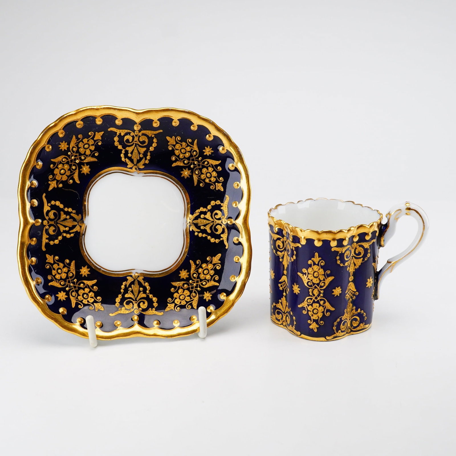 Coalport Dark Blue and Gold Cup and Saucer Set: This porcelain demitasse cup and saucer set is produced by Coalport and finished in a deep cobalt blue glaze with elaborate raised gold paste decoration. Both pieces feature a distinctive quatrefoil o