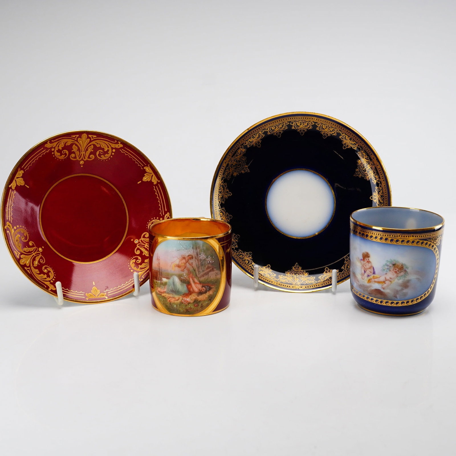 Set of Two Hand-Painted Porcelain Cabinet Cups and Saucers, Haviland and Royal Vienna: This lot consists of two sets of hand-painted porcelain cabinet cups and saucers, totaling four pieces, featuring cherub and mythological themes. The first set is finished in a rich claret red ground 