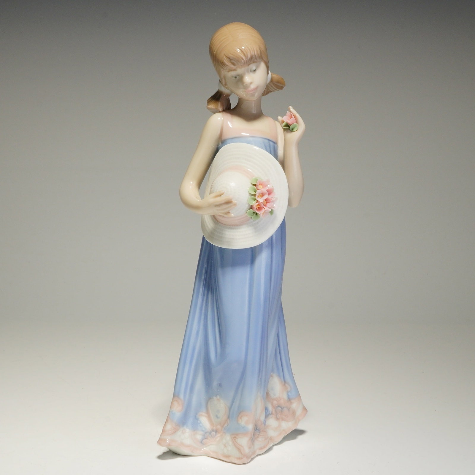 Lladro Porcelain Figurine, Courtney 01005648: The piece depicts a young girl with pigtails wearing a long, light blue sleeveless dress with a floral-patterned hem. She is posed holding a single pink blossom in one hand and a white wide-brimmed