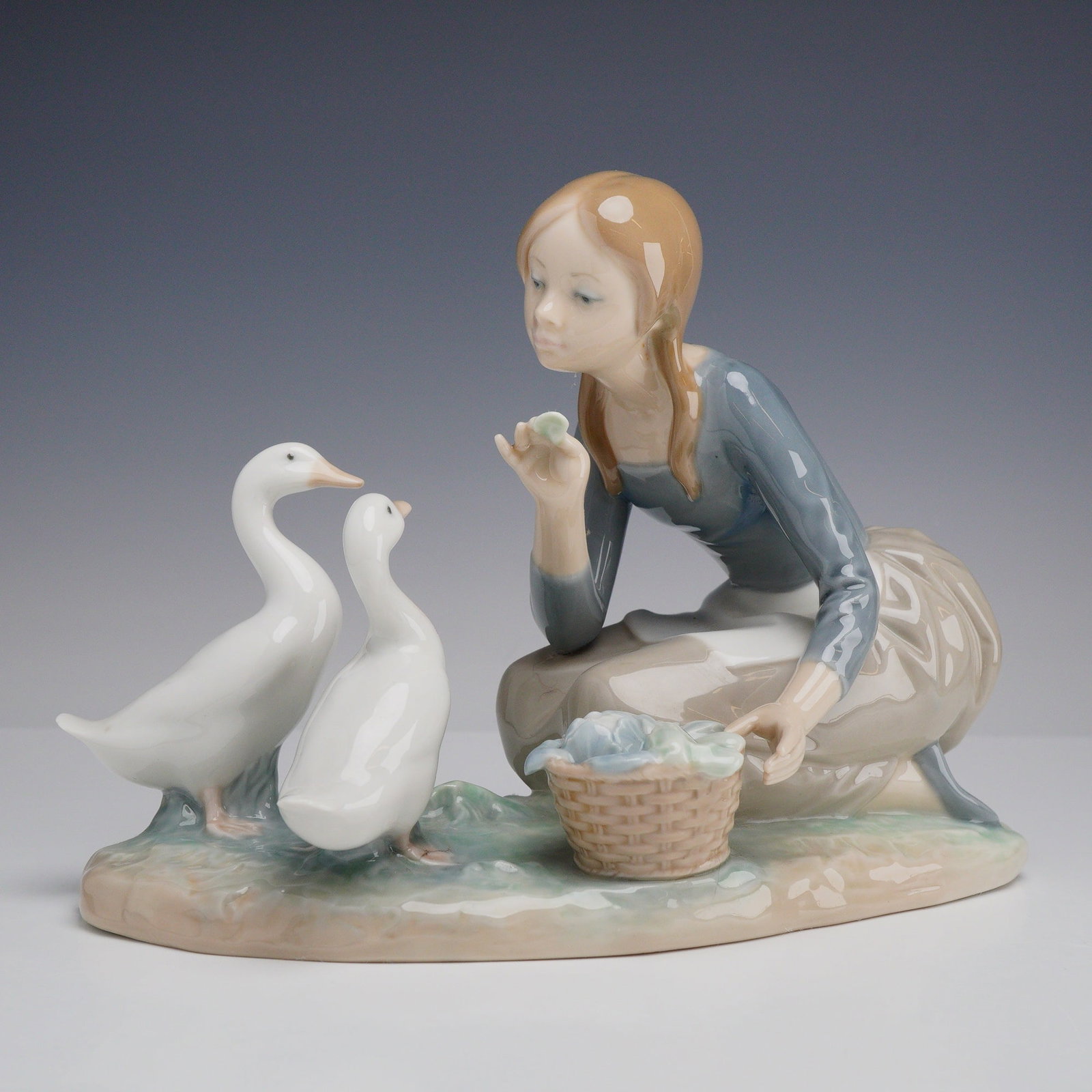 Lladro Porcelain Figurine Feeding the Ducks 01004849: The piece features a young girl in a kneeling pose, wearing a blue long-sleeved bodice, a tan skirt, and a white apron. She is shown gesturing toward two white ducks positioned on a grassy base. A sma