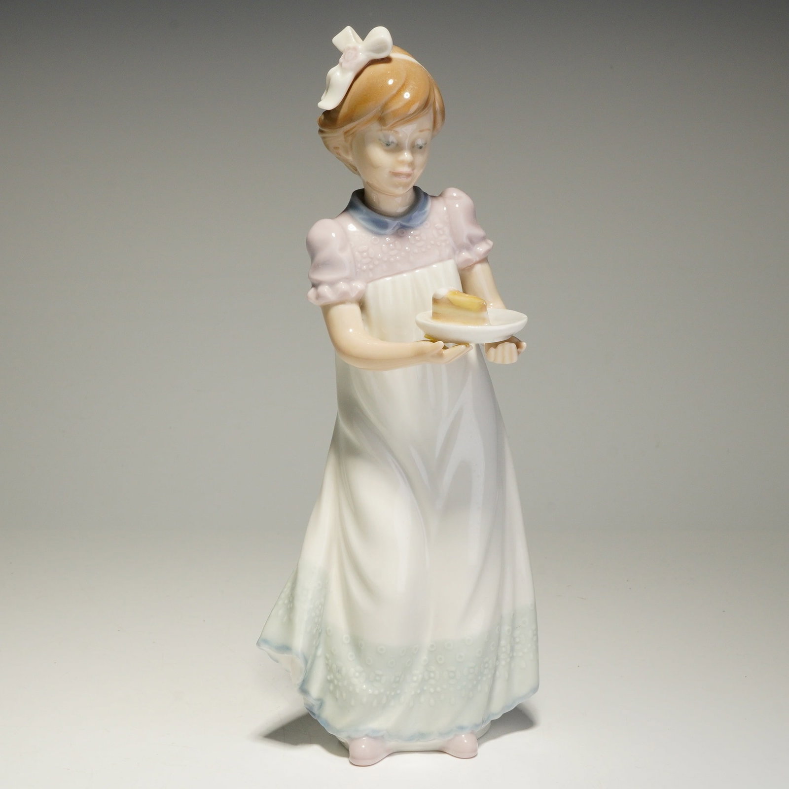 Lladro Porcelain Figurine, Happy Birthday 01005429: The figure depicts a young girl standing and holding a small white plate with a single slice of cake. She is dressed in a full-length white nightgown with a pale pink bodice, a blue collar, and a ligh