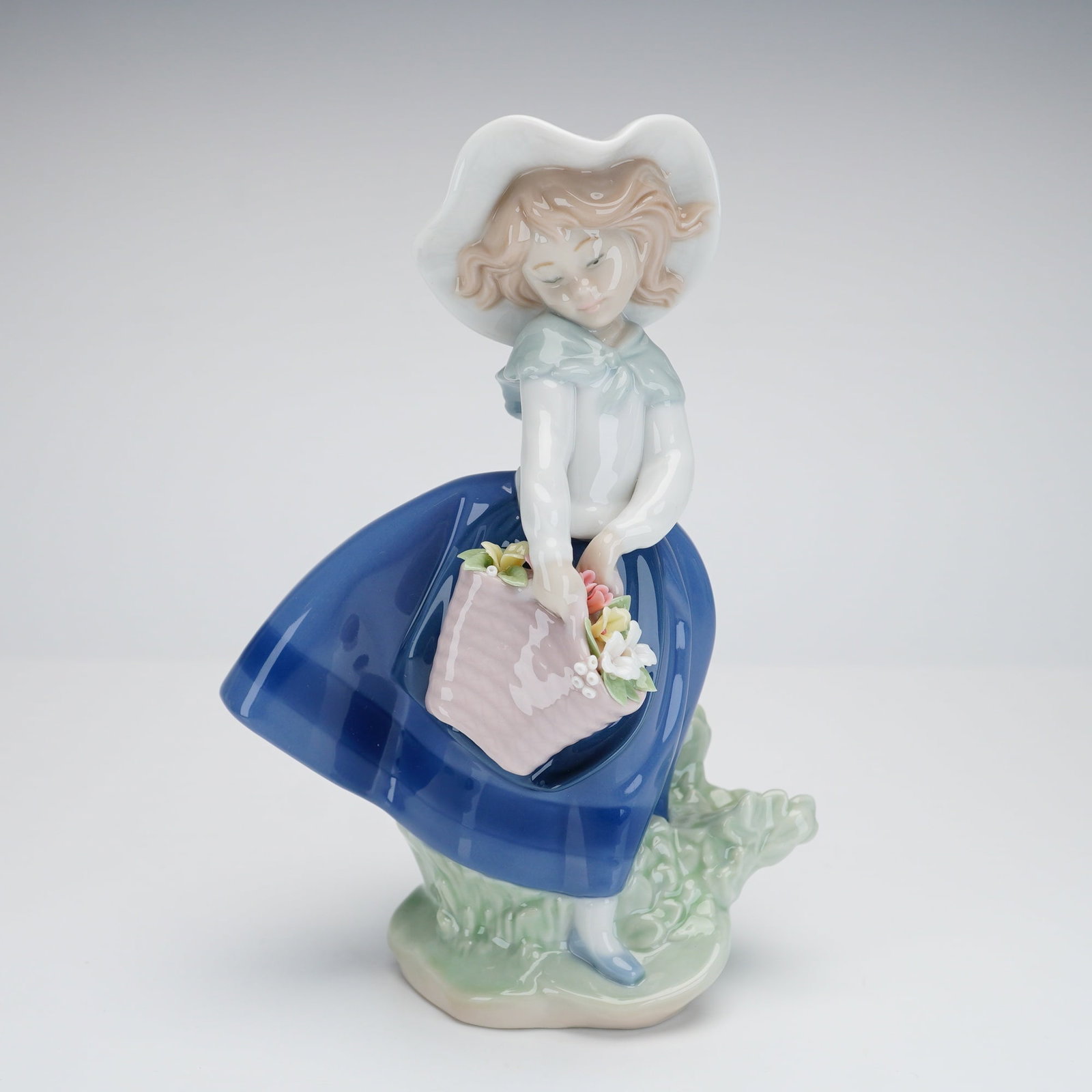 Lladro Pretty Pickings Porcelain Figurine 1005222: This Lladro glazed porcelain figurine, titled Pretty Pickings and identified as model number 5222, depicts a young girl in a wide-brimmed white hat, light blue blouse, and a flowing deep blue skirt. T