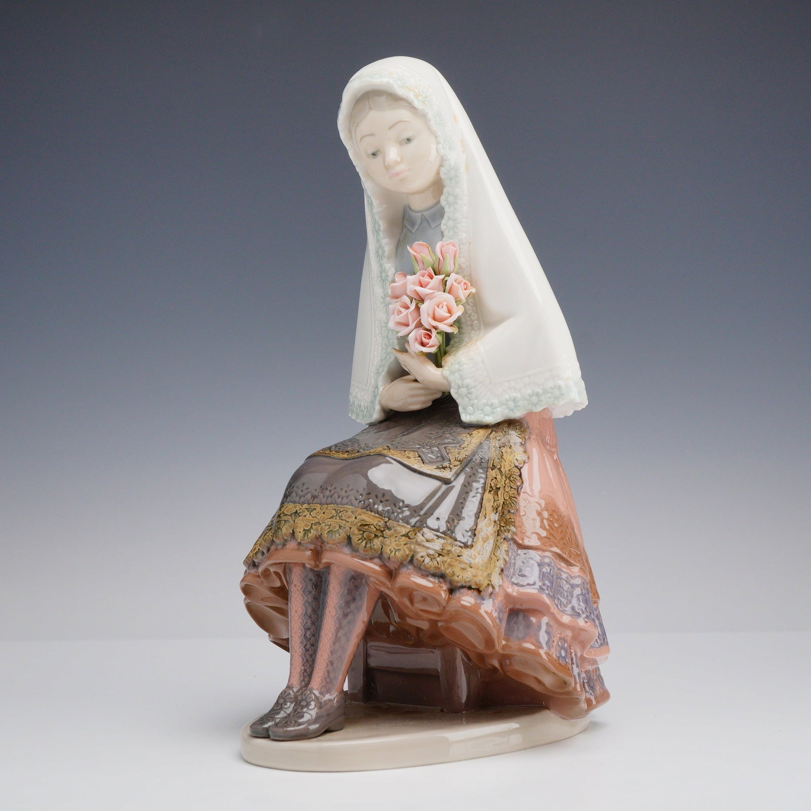 Lladro Porcelain Figurine Girl Sitting with Roses 01005127: This Lladro glazed porcelain figurine depicts a young woman in traditional Spanish Valencian attire, seated on a low brown stool in a contemplative pose. The figure wears a white lace-trimmed mantilla