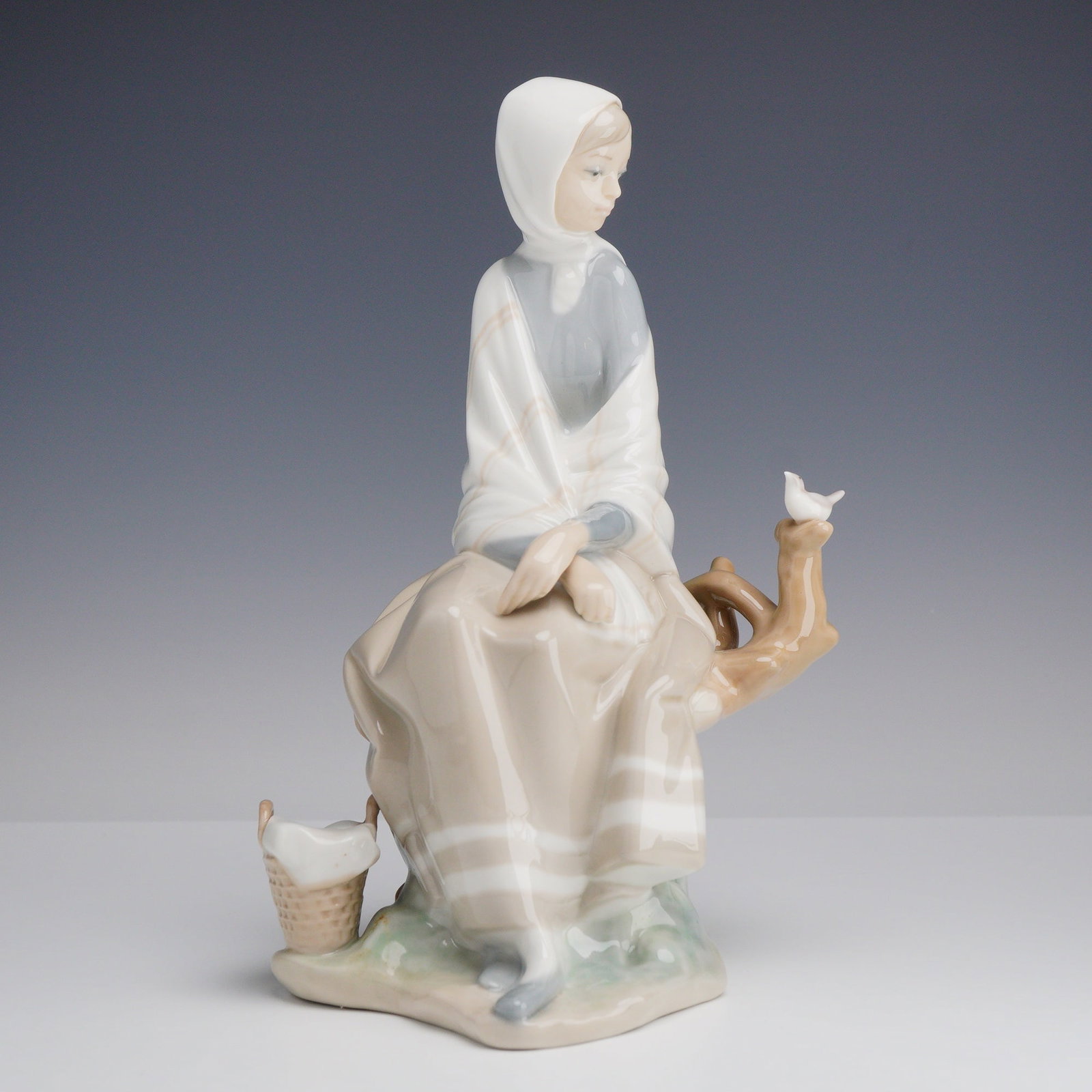 Lladro Porcelain Figurine New Shepherdess 01004576: This Lladro porcelain figurine depicts a young woman in a pensive, seated pose atop a stylized tree branch. The figure is dressed in a white headscarf and a gray shawl draped over a long, pale tan dre