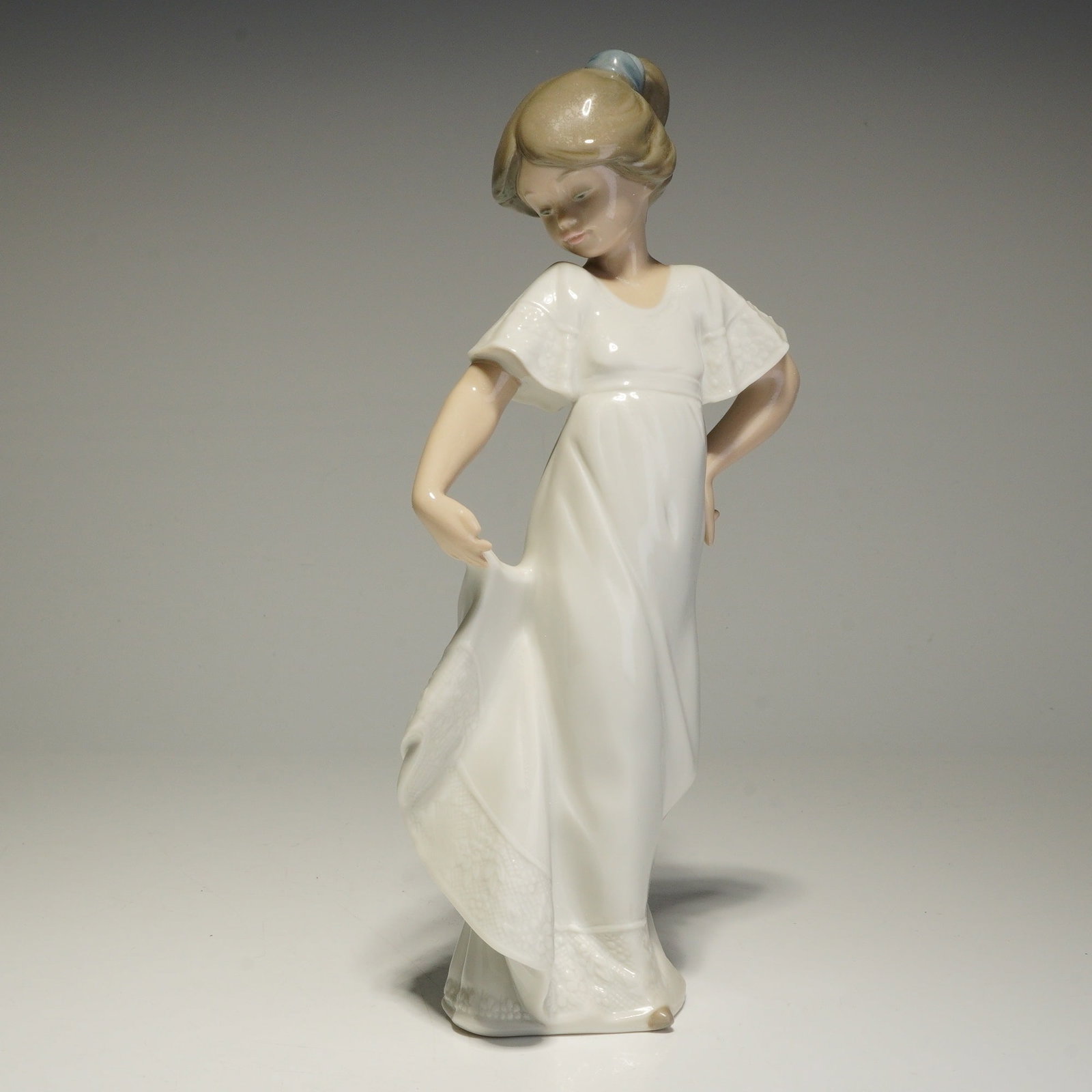 Nao by Lladro Porcelain Figurine, How Pretty 02001110: The figure depicts a young girl in a long white dress holding the side of her skirt. The piece is finished in a glazed pastel palette with a blue hair bow and molded lace-like patterns on the sleeves 