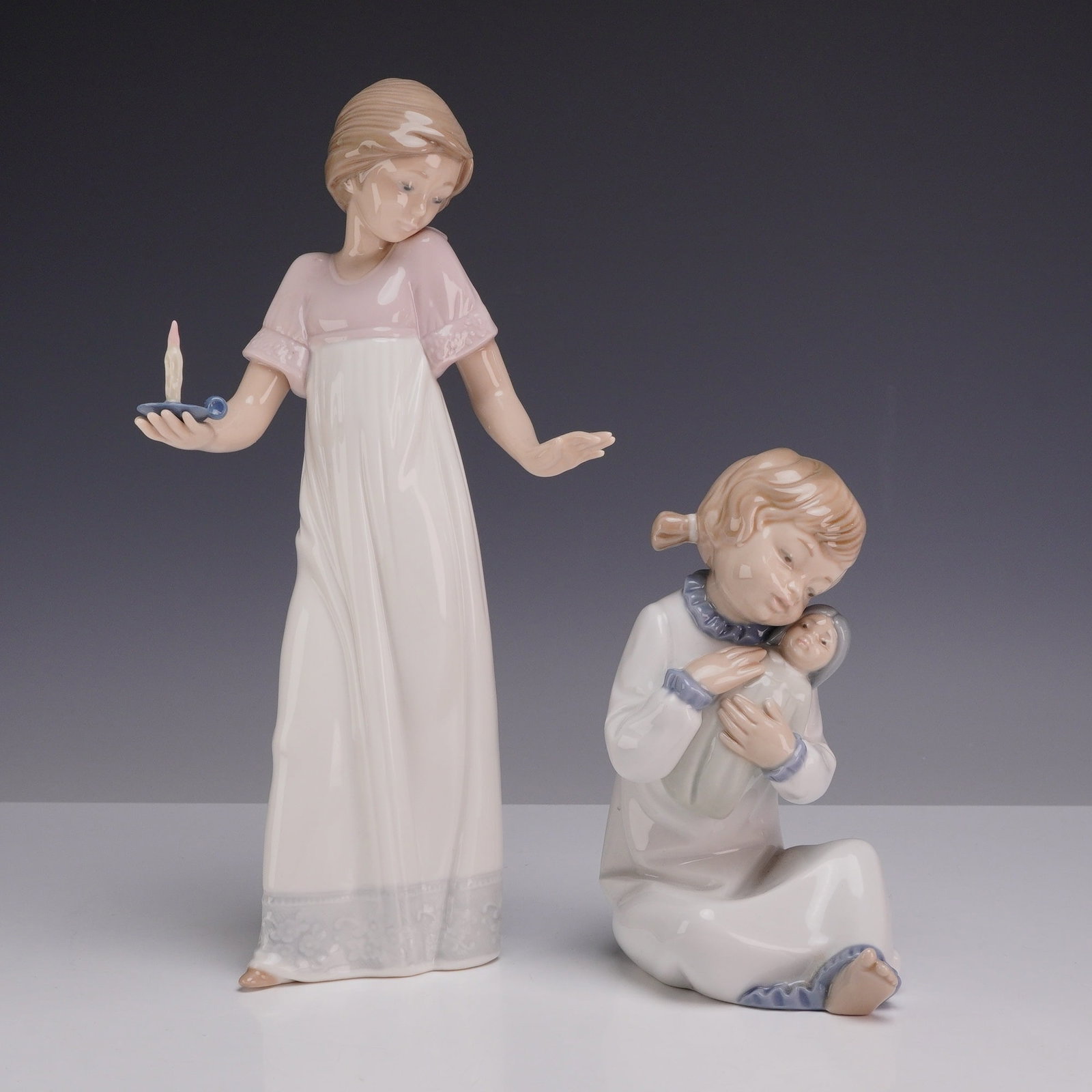 2pc Nao by Lladro Porcelain Figurines of Young Girls in Nightwear: A pair of glazed porcelain figurines manufactured by Nao, a member of the Lladro Group, produced in Spain. Both figures depict young girls dressed in nighttime attire. The first figurine features a st