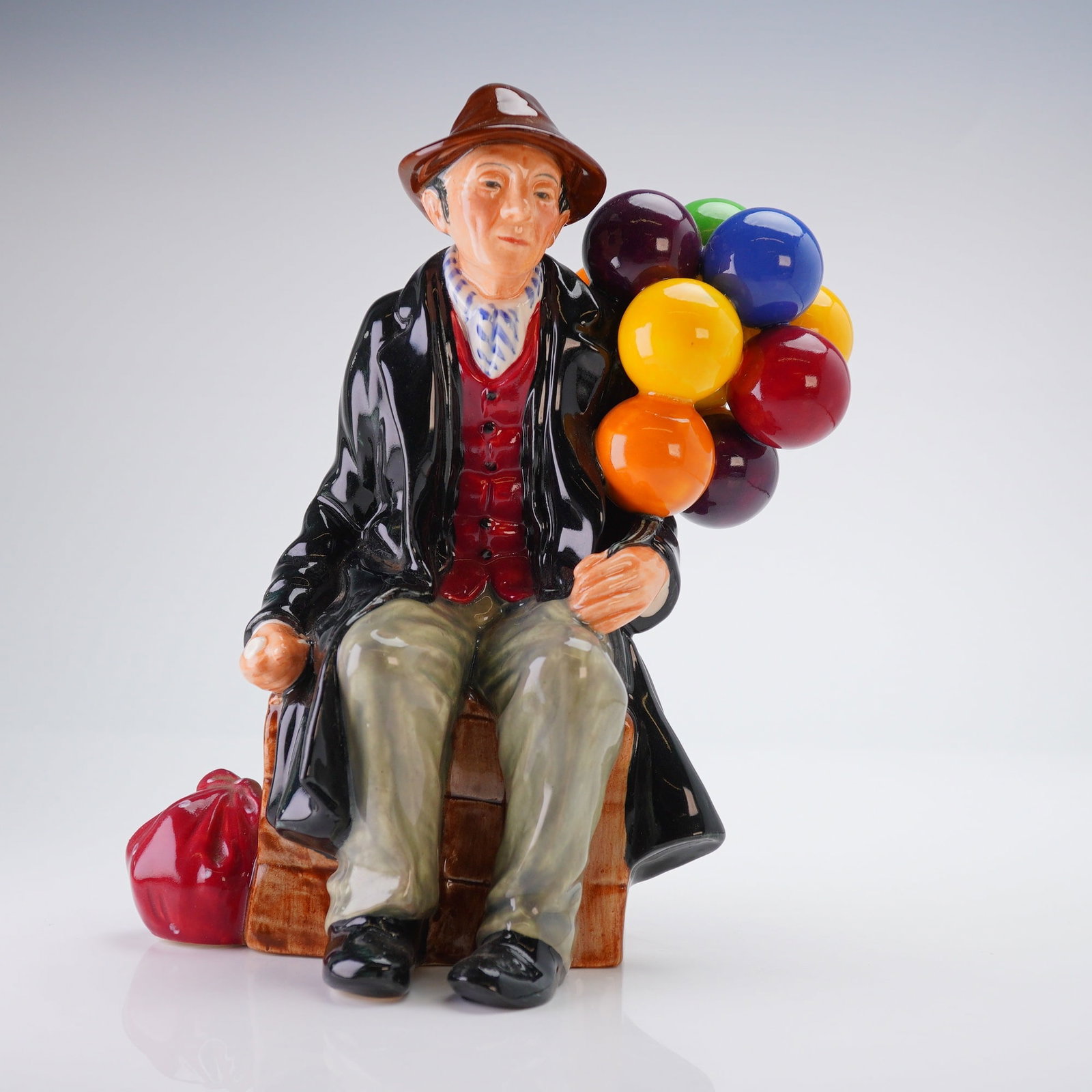 Royal Doulton Figurine The Balloon Man HN1954: This polychrome figure depicts an elderly man wearing a brown hat and a long black overcoat over a red waistcoat. The subject is seated on a textured crate and holds a cluster of multicolored balloons