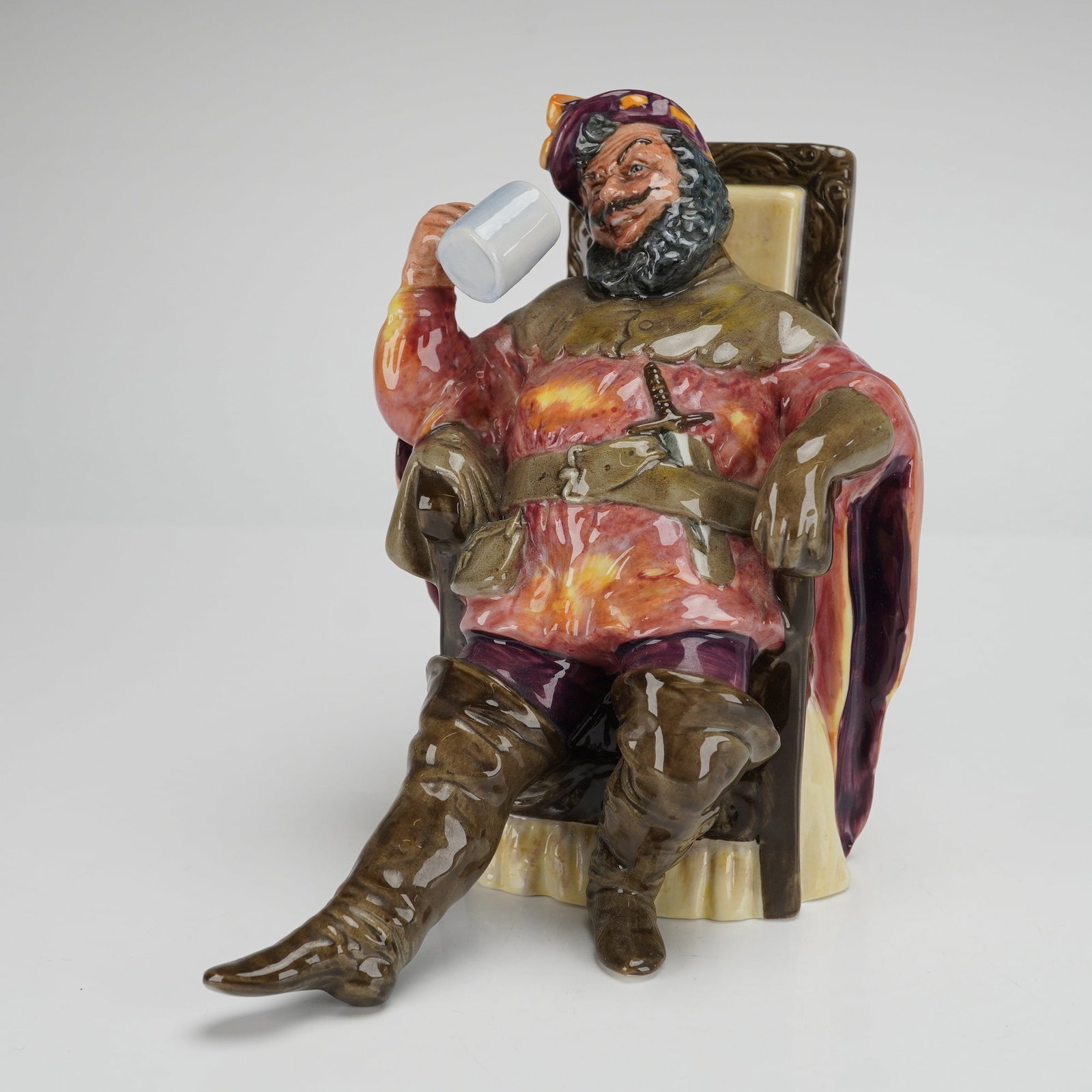 Royal Doulton Figurine The Foaming Quart HN2162: The figure depicts a seated man in colorful period costume, holding a white drinking mug to his mouth. The figure is modeled in highly saturated, high-gloss polychrome glazes, featuring deep browns, r