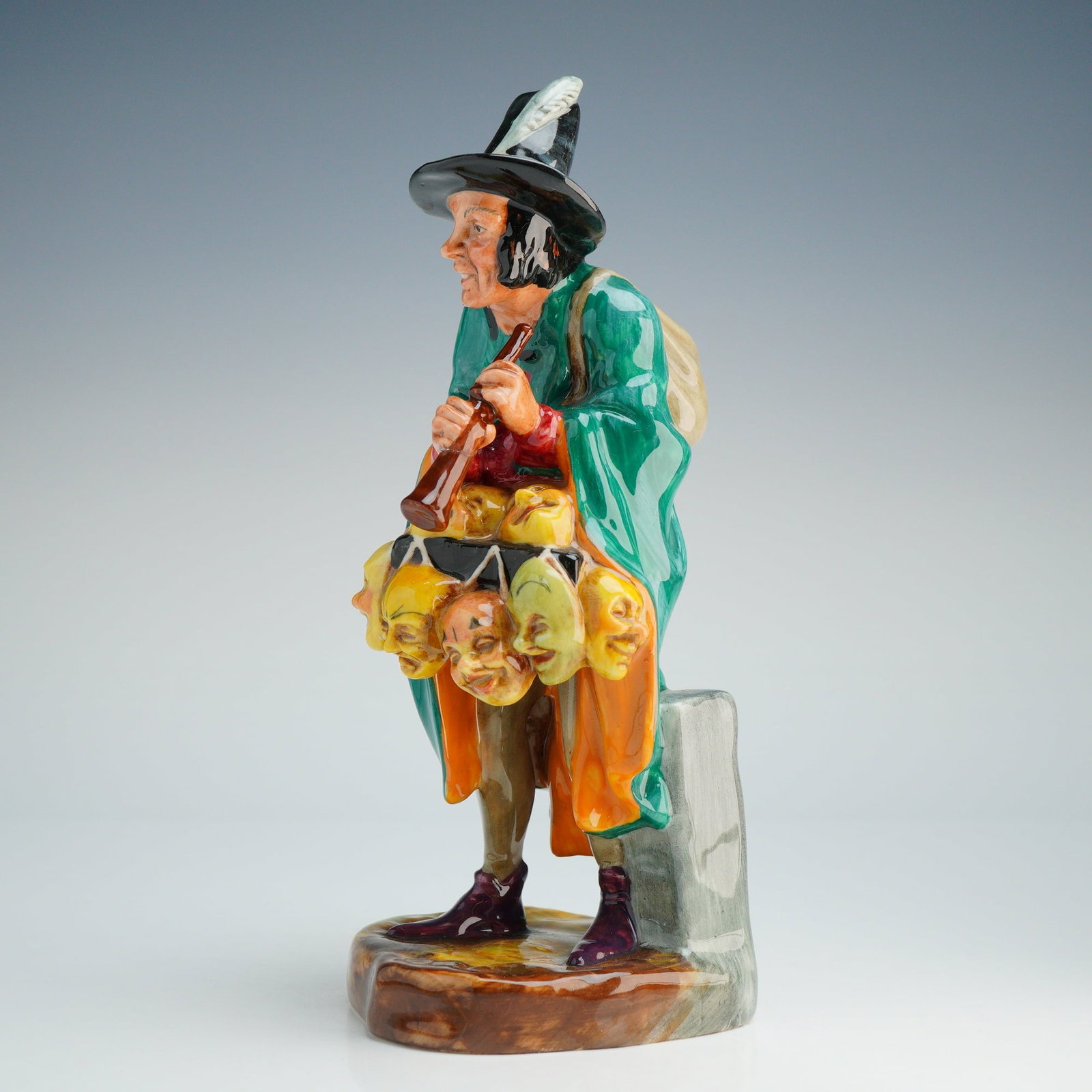 Royal Doulton Figurine The Mask Seller HN2103: This Royal Doulton depicts a wandering merchant in a green cloak and a tall black hat accented with a white feather. The figure is modeled playing a flute with a tan sack on his back and a lantern han