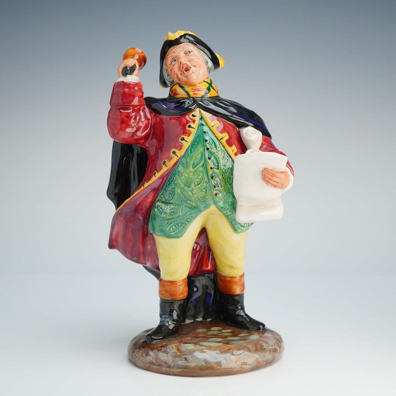 Royal Doulton Figurine Town Crier HN2119: A Royal Doulton designed by Peggy Davies depicts a male town crier in traditional mid-18th-century attire, modeled in a dynamic pose with his mouth open as if shouting. He holds a handbell in his rais