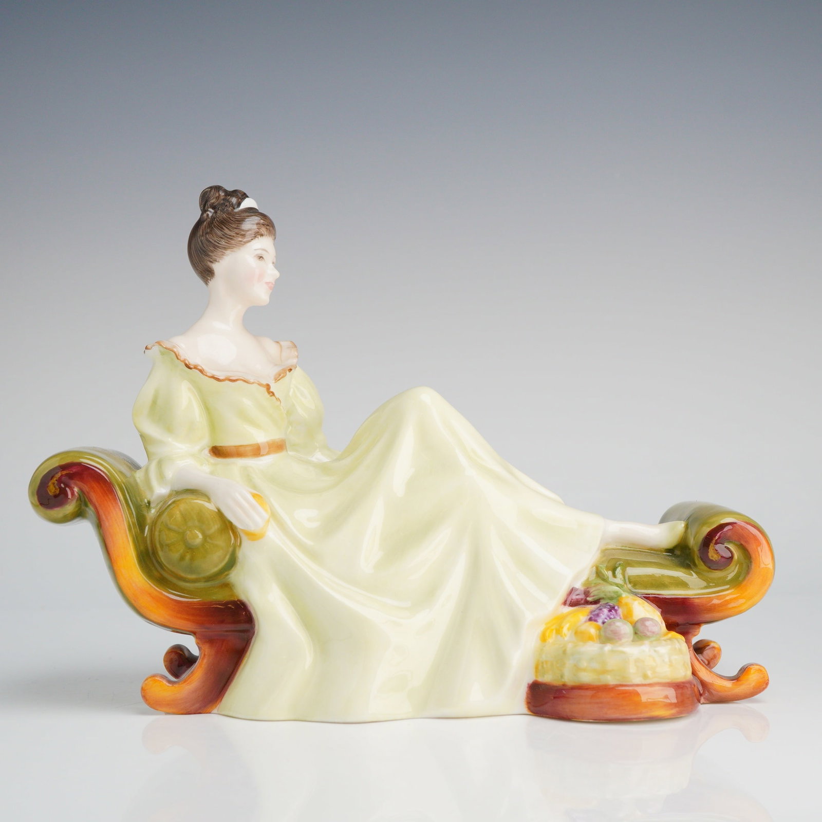 Royal Doulton Figurine, At Ease HN2473: The piece is crafted from glazed ceramic, likely porcelain or bone china, depicting a woman in a historical style gown reclining upon a chaise longue. The woman wears a pale-yellow dress with a tan sa
