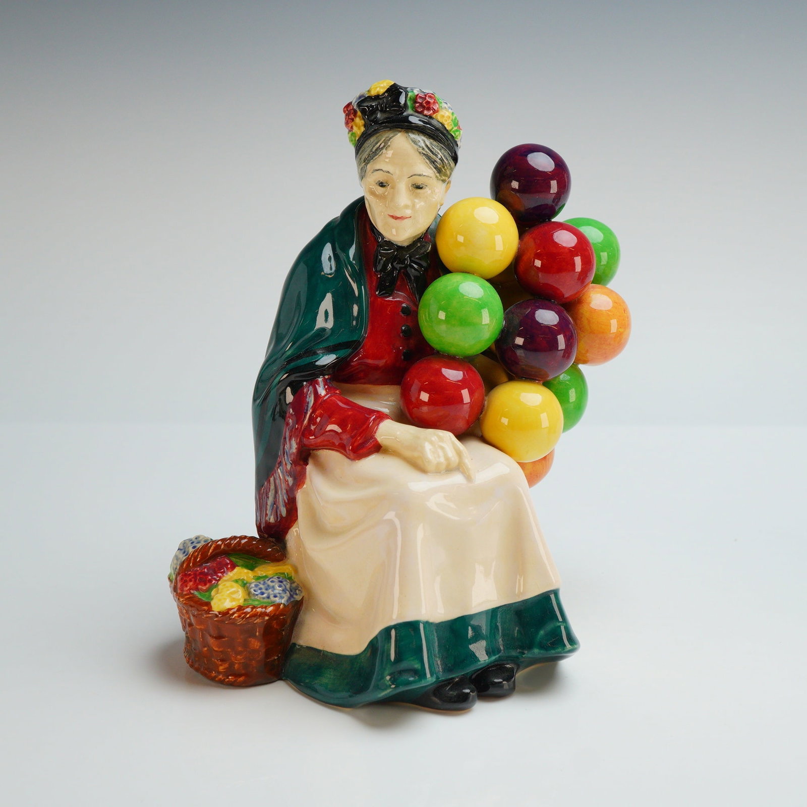 Royal Doulton Figurine, Old Balloon Seller HN1315: The figure depicts an elderly woman seated on a brown crate. She is dressed in a green shawl over a red bodice and a white apron with a dark green border, with a patterned headscarf tied over her grey