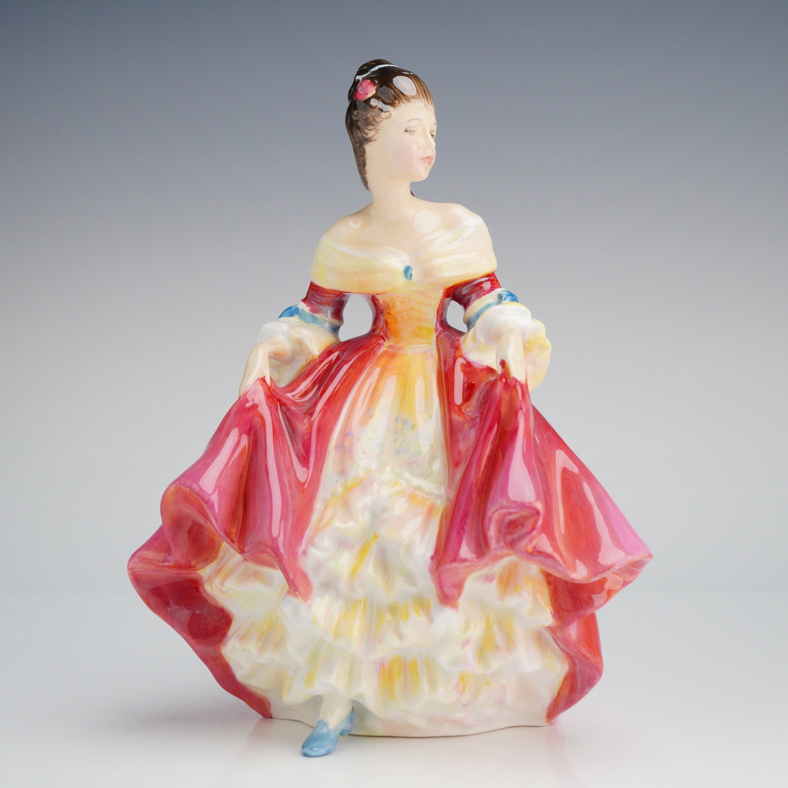 Royal Doulton Figurine, Southern Belle HN2229: The figure is depicted wearing a dramatic historical style gown, finished in high-gloss polychrome enamels dominated by deep red, yellow, and white ruffles, accented with subtle blue details on the bo