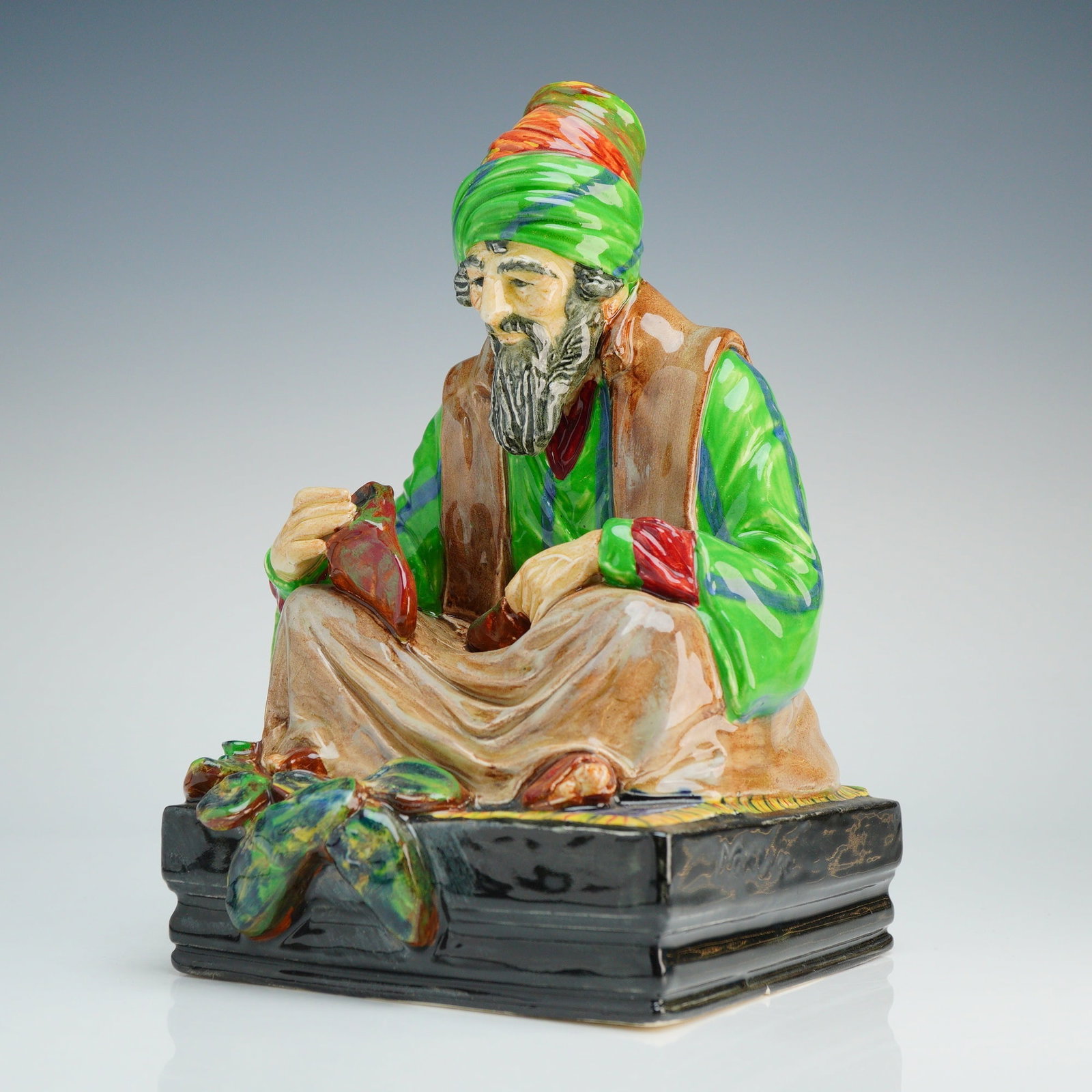 Royal Doulton Figurine, The Cobbler HN1706: Royal Doultondesigned by Charles John Noke depicts a bearded man in Middle Eastern-style attire, including a green robe and a green and orange turban. The subject is seated cross-legged on a colorful 