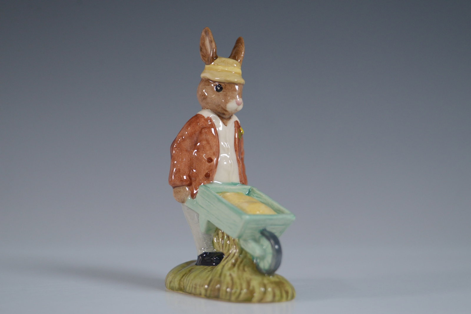 Royal Doulton Bunnykins Figurine, Gardener Bunnykins DB156: Charming Royal Doulton Bunnykins figurine titled Gardener Bunnykins, model DB156. This delightful ceramic figure features a rabbit in a brown jacket and yellow hat, pushing a green wheelbarrow filled 