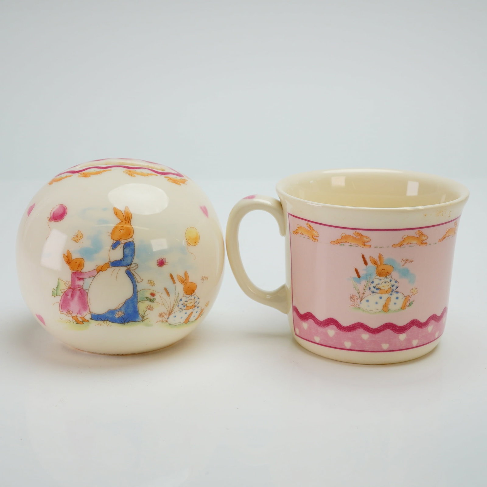 Two Royal Doulton Bunnykins Bone China Money Ball and Mug: Two pieces of Royal Doulton Bunnykins nursery ware in the Sweetheart H5317 pattern. The set is crafted from fine bone china and features a cream-colored glaze decorated with colorful transfer-printed 