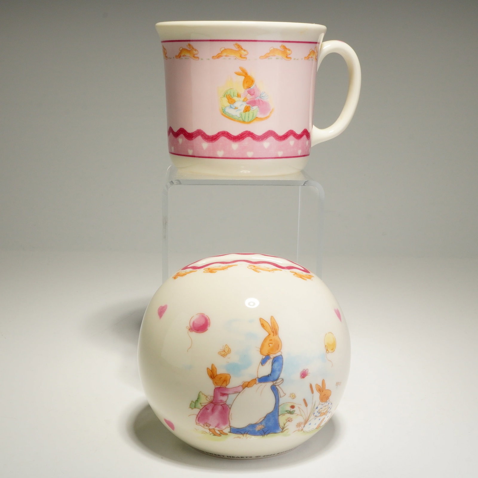 2pc Royal Doulton Bunnykins Sweet Hearts Mug and Money Bank: The handled mug and spherical money box are crafted from bone china with a cream-colored ground, while the mug features a prominent pink area as its main background. Both pieces showcase colorful illu
