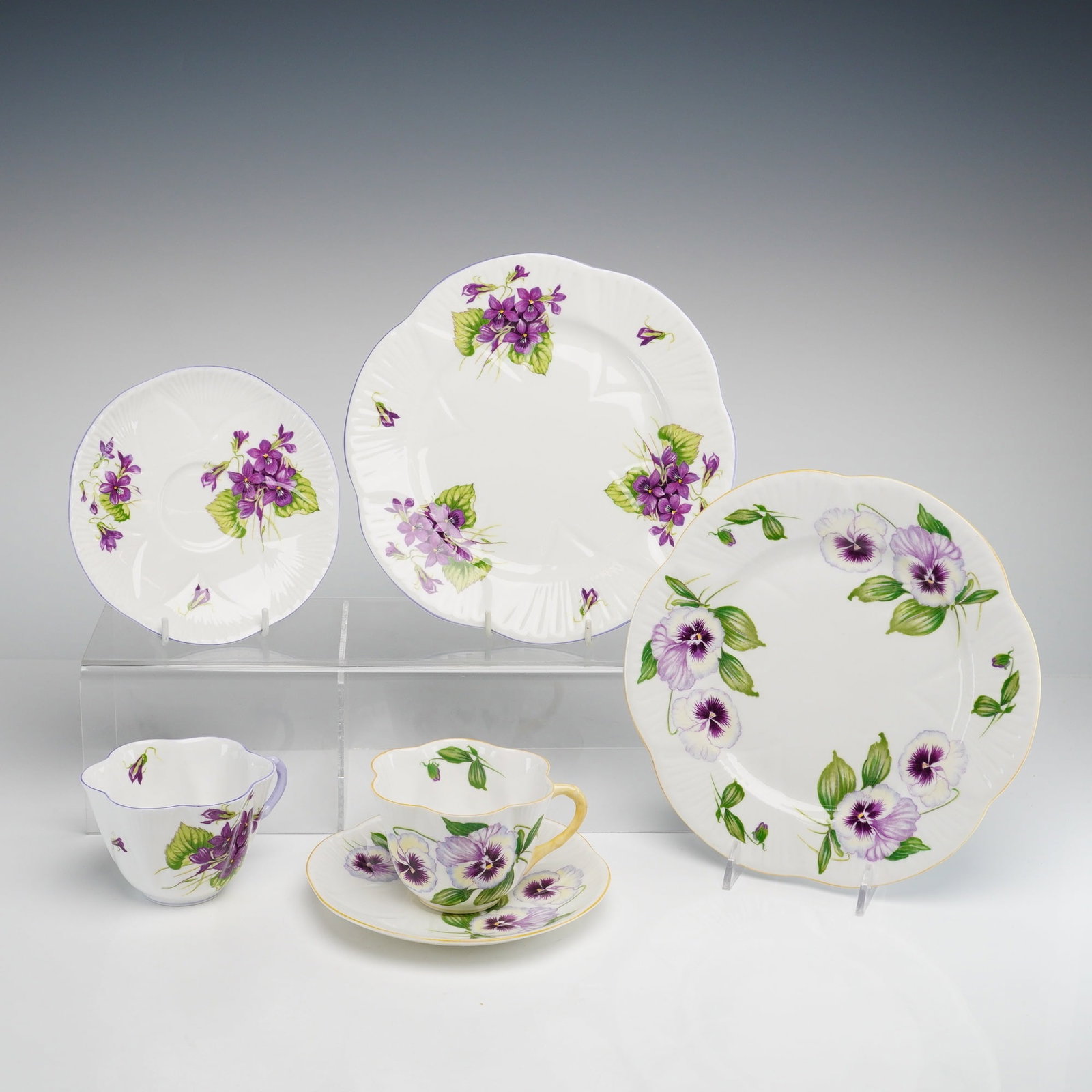Lot of Two Shelley Bone China Cup, Saucer and Dish Sets, Violets: Each trio includes three pieces: a teacup, a saucer, and a dessert plate. All pieces are modeled in the Dainty shape, which features deeply lobed and scalloped rims. The first trio is decorated in the