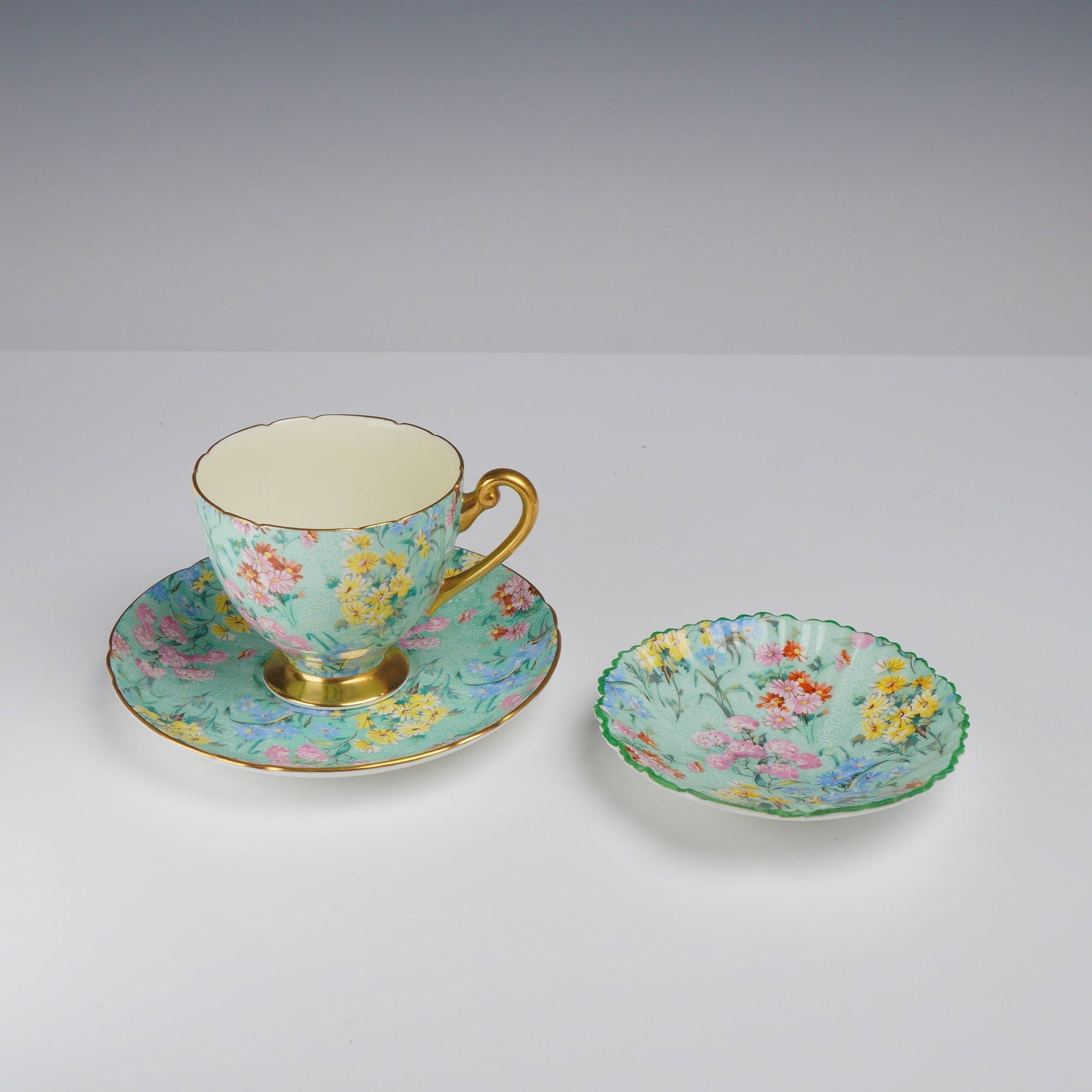 3pc Shelley Bone China Tea Trio, Melody: This lot features three pieces of Shelley fine bone china in the Melody chintz pattern, manufactured in England. The group includes a footed teacup with an ornate handle, a coordinating saucer, and a 