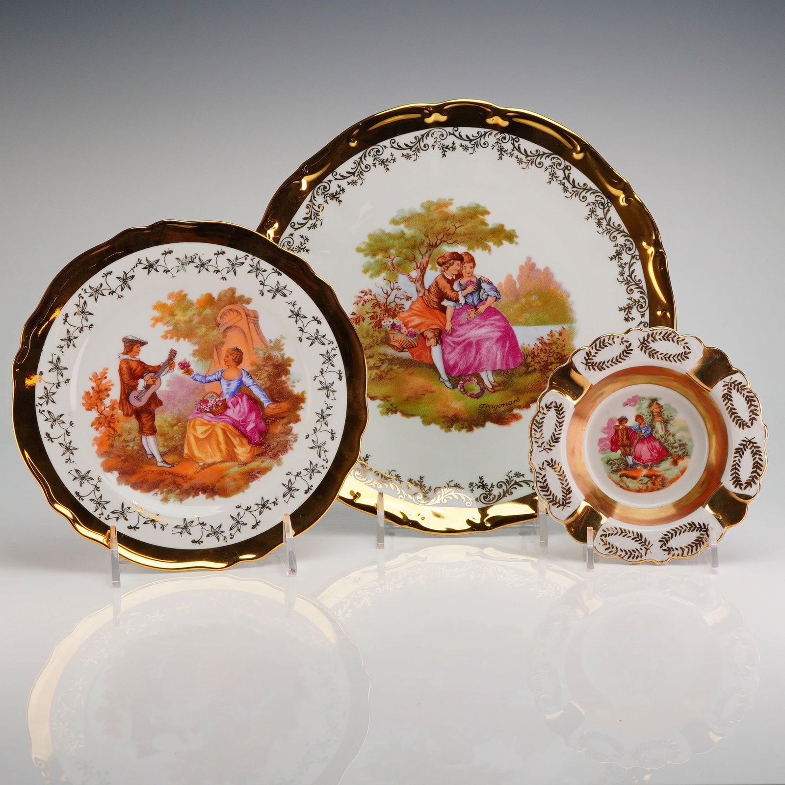 3pc German Porcelain Plates and Ashtray, Fragonard Courting Scene: The set is executed in a Rococo revival style, featuring scalloped, undulating rims and extensive 22 karat gold gilding (Goldauflage) applied over white porcelain. The central decoration on each item 