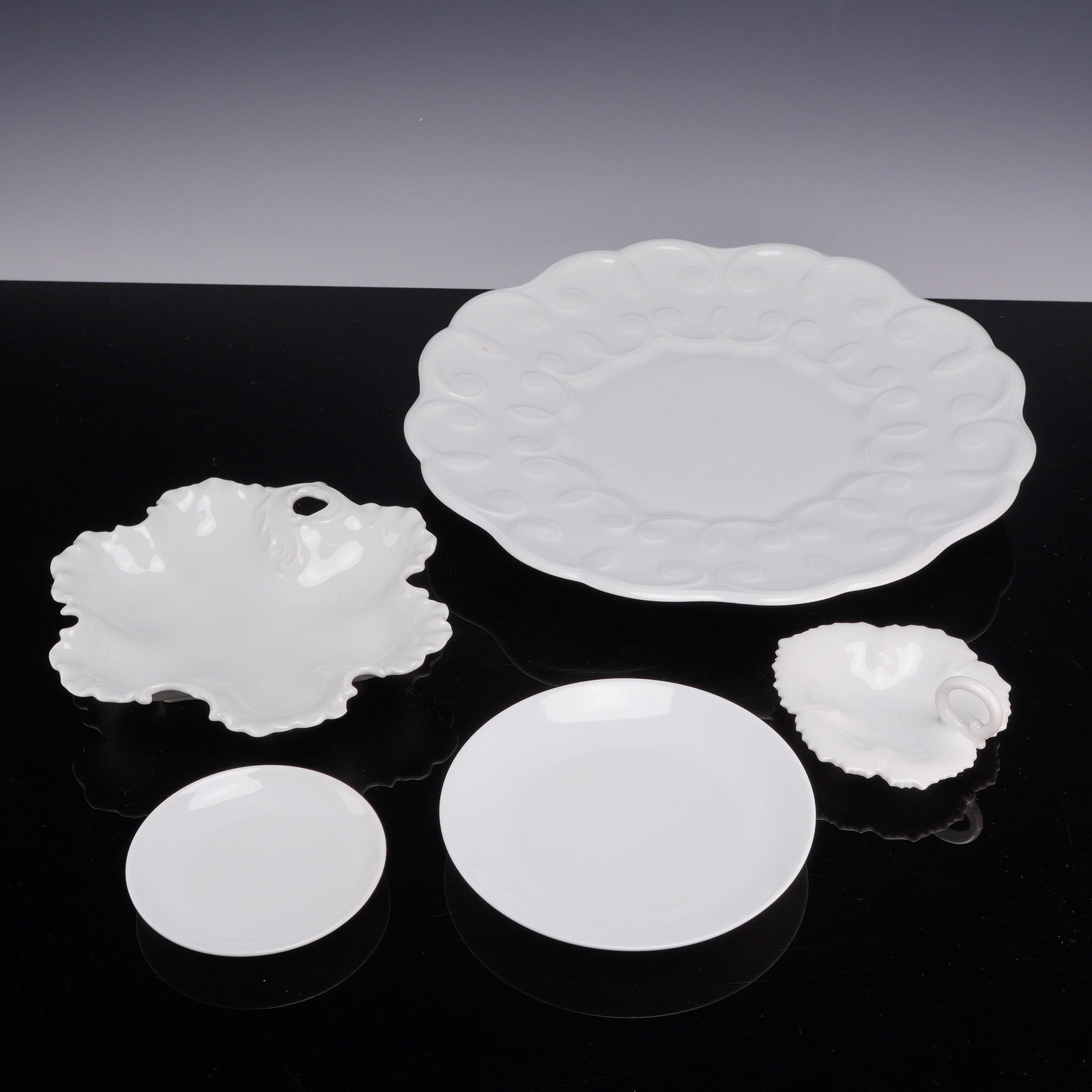 A Collection of Five White Porcelain Serving Pieces including J.L. Coquet: The group includes a large circular platter featuring a scalloped rim and a wide border decorated with an embossed interlocking scroll and loop motif. Also included in the collection is an organic lea