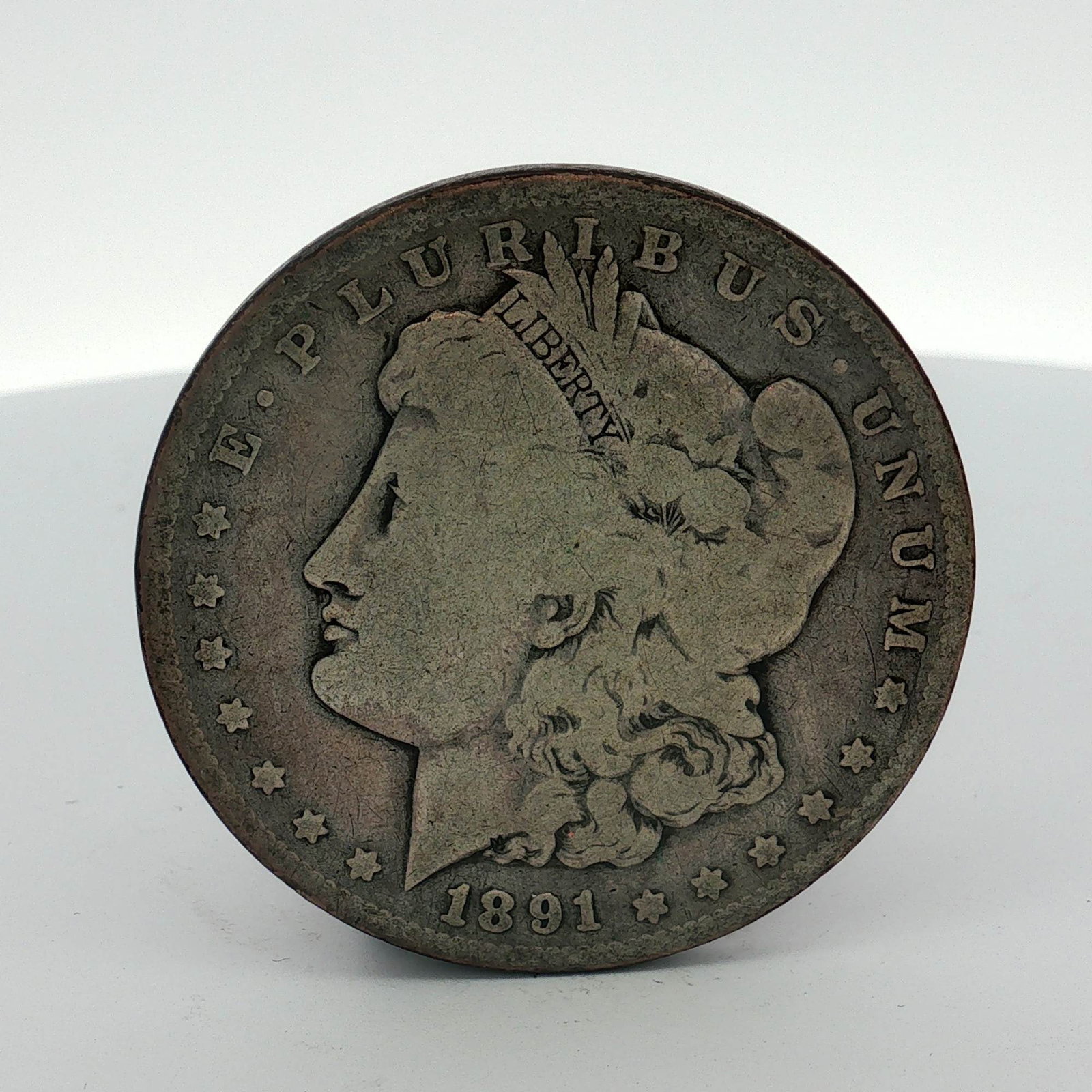 1891 US Morgan Silver Dollar Coin Circulated: This US silver coin is a Morgan dollar struck at the Philadelphia Mint. The obverse features the Liberty head design and is dated 1891. The reverse displays the no mint mark located below the wreath a