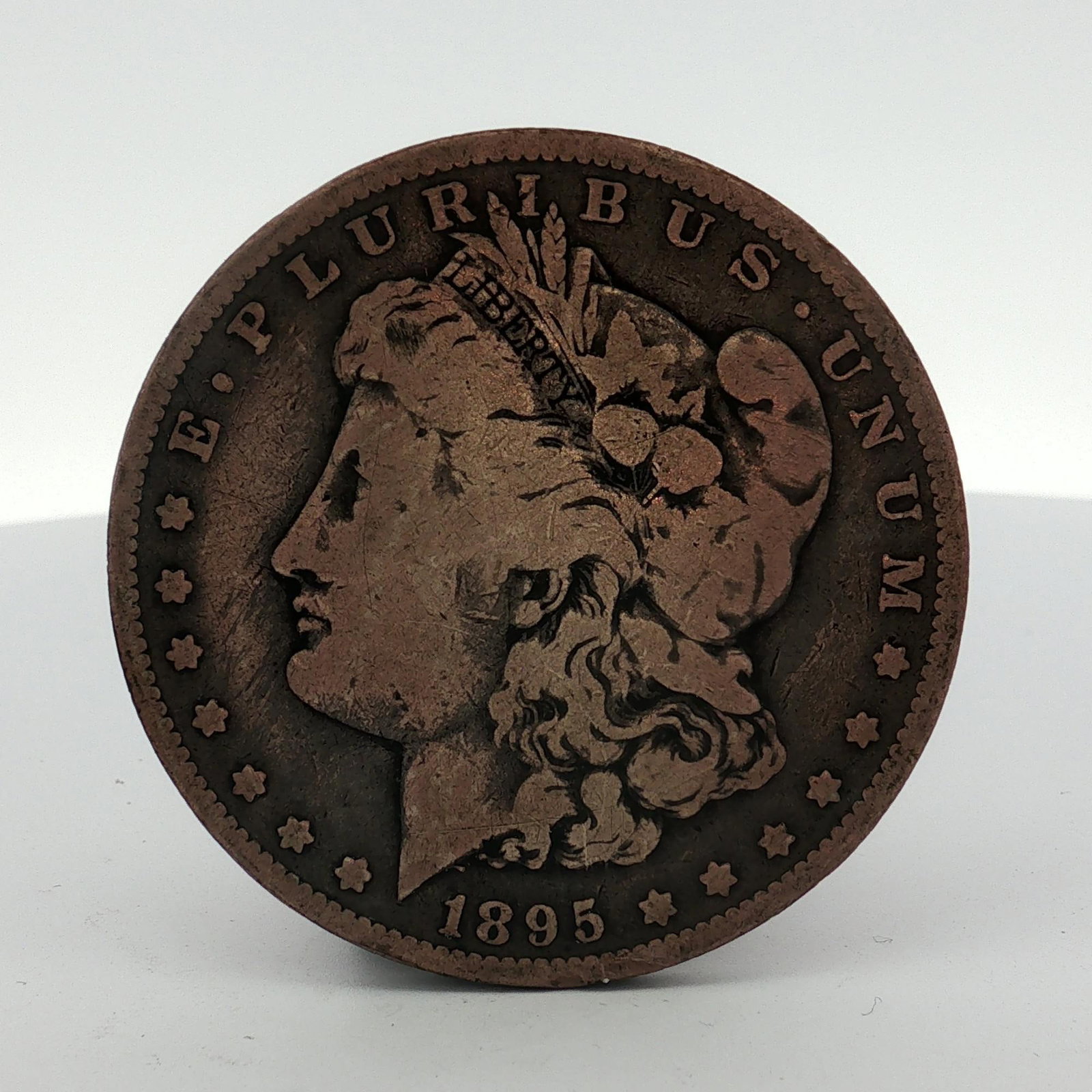 1895-O US Morgan Dollar: This United States Morgan dollar coin is dated 1895 and features the New Orleans "O" mint mark. The specimen displays a distinctive dark, copper-toned surface and a granular, matte texture throughout.