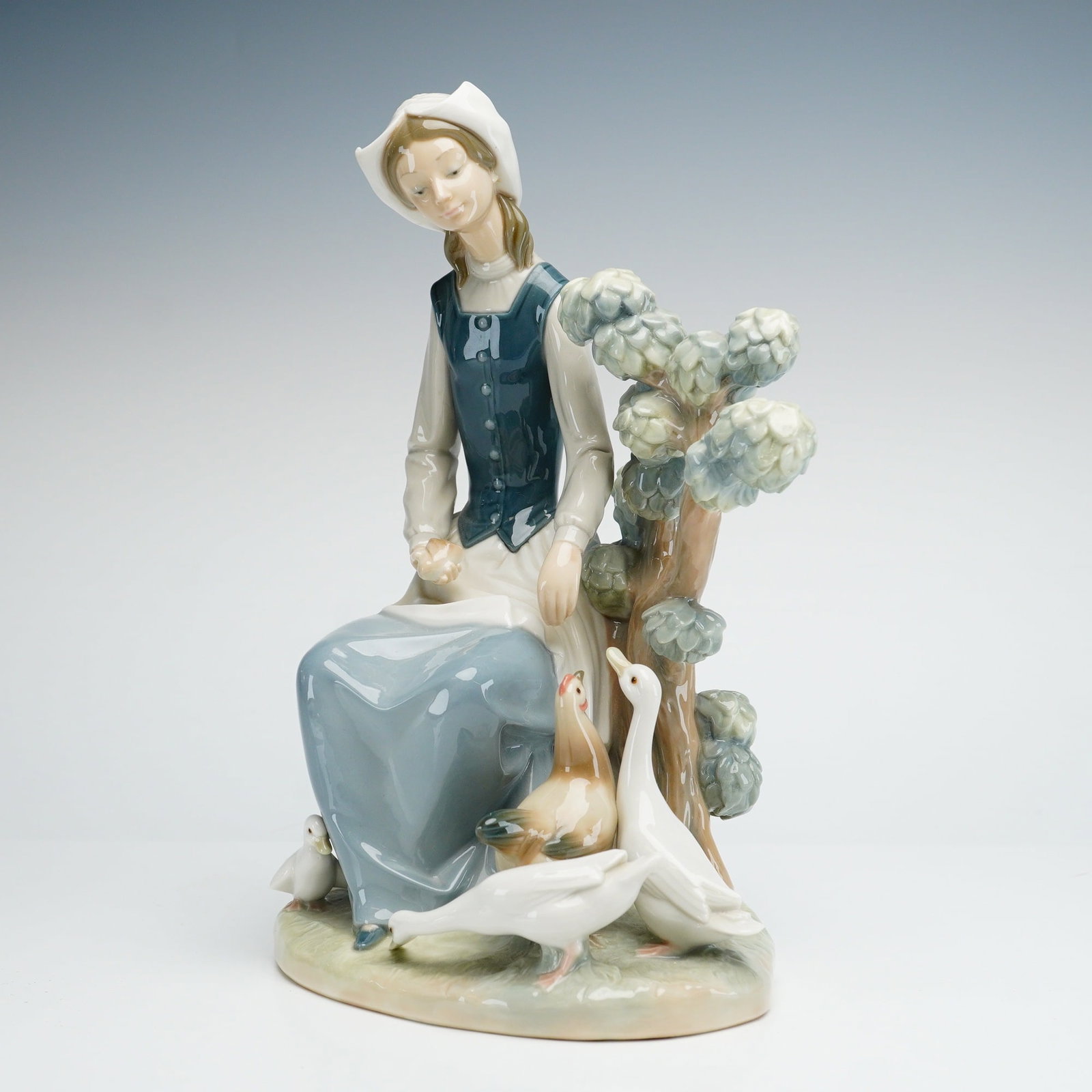 Lladro Glazed Porcelain Figurine Seated Young Woman with Geese: This is a glazed porcelain figurine from the Spanish manufacturer Lladro. The subject depicts a young woman in traditional rural attire, including a blue vest over a white long-sleeved blouse, a grey-