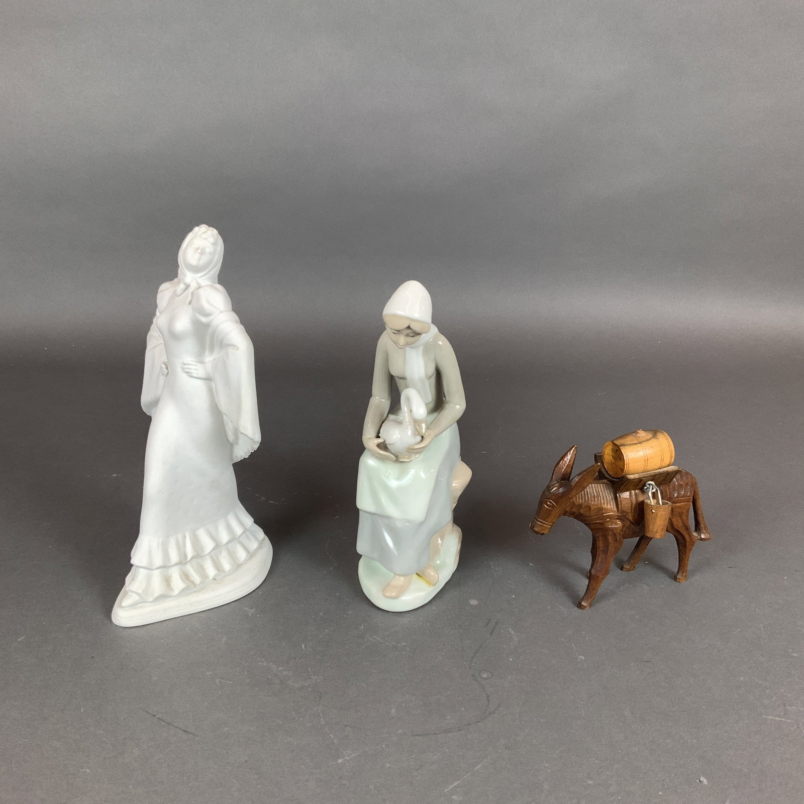 3pc Porcelain and Wood Figurines: This collection includes three decorative figurines. The first is a standing Spanish woman figurine made of white bisque porcelain. The figure is draped in a shawl and traditional dress. The underside