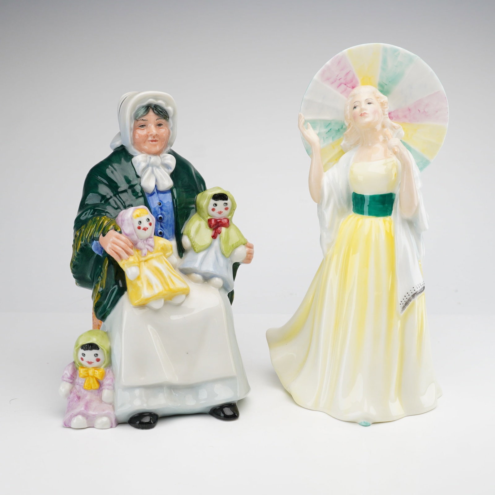 2pc Royal Doulton Figurines, The Rag Doll Seller HN2944 and Jane HN2806: This lot contains two Royal Doulton glazed bone china figurines, "The Rag Doll Seller" and "Jane." The Rag Doll Seller (HN 2944) depicts an elderly woman seated on a woven wicker basket. She is dresse