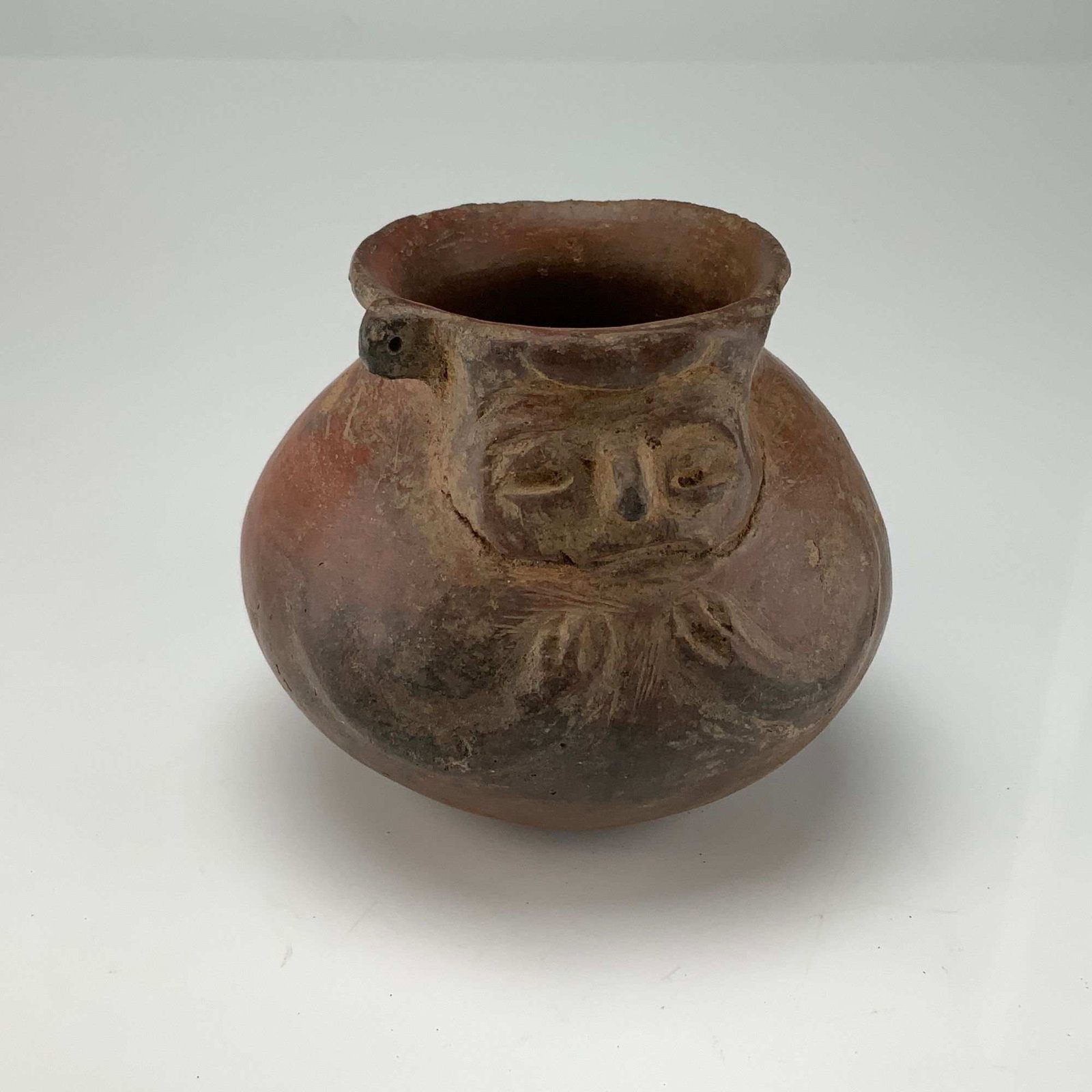 Pre-Columbian Style Ceramic Face-Neck Vessel: This ceramic effigy vessel is an example of Pre-Columbian style pottery. The object is a small face-neck jar, characterized by a stylized human face modeled onto the neck section just below the rim.