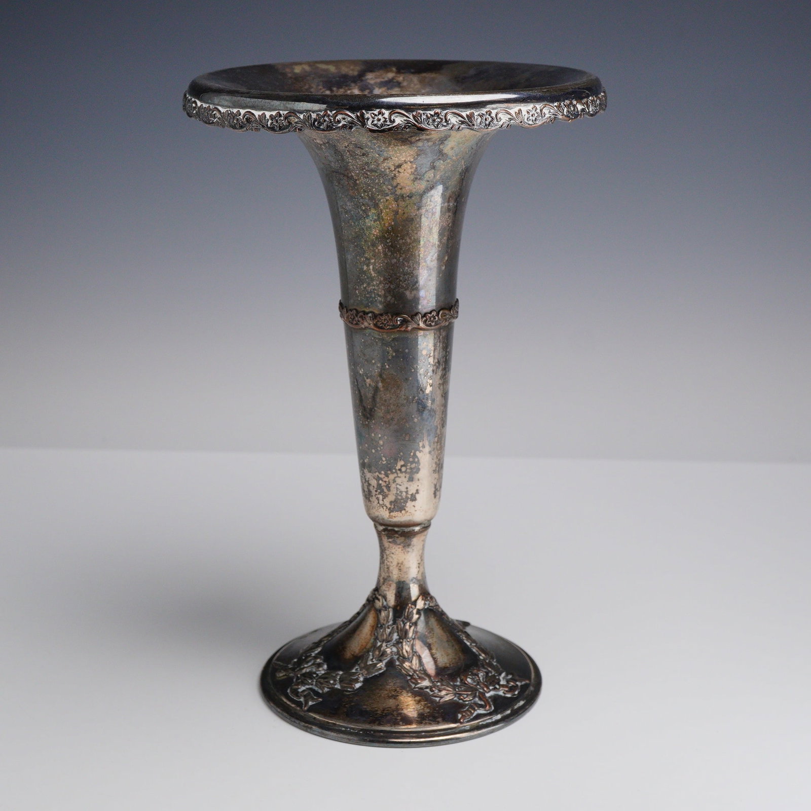 Silver Plated Trumpet Vase: This silver-plated trumpet vase features a widely flared rim with a cast foliate and floral border. The tapering cylindrical body is embellished with a central decorative band containing matching
