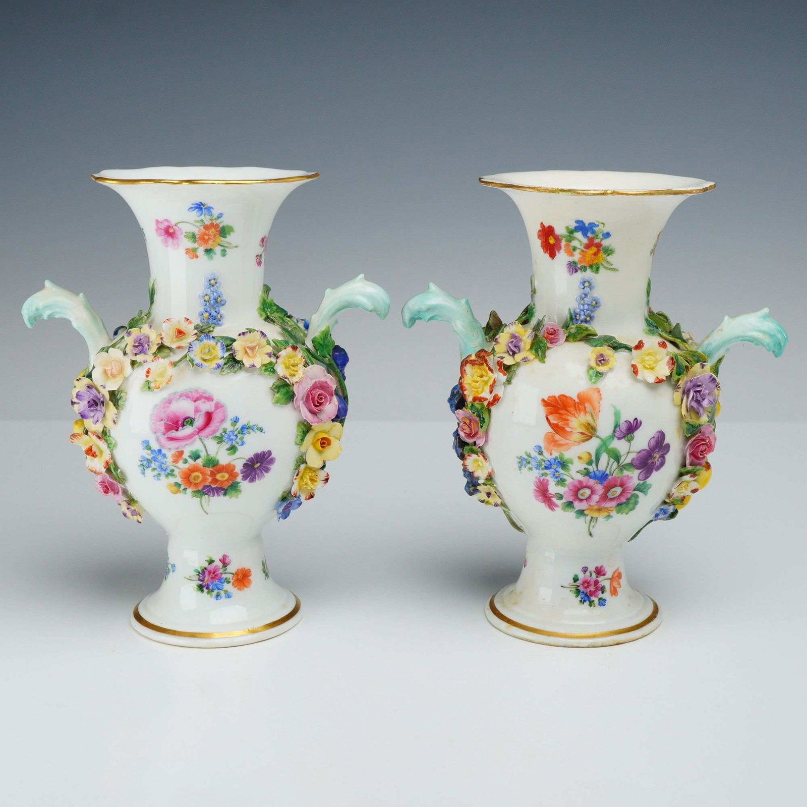 Pair of Meissen Encrusted Floral Porcelain Vases: A pair of Meissen porcelain twin-handled vases. Each vase is designed in the Rococo revival style, featuring a flared, scalloped rim with a gilded edge and a tapered neck. The ovoid bodies are
