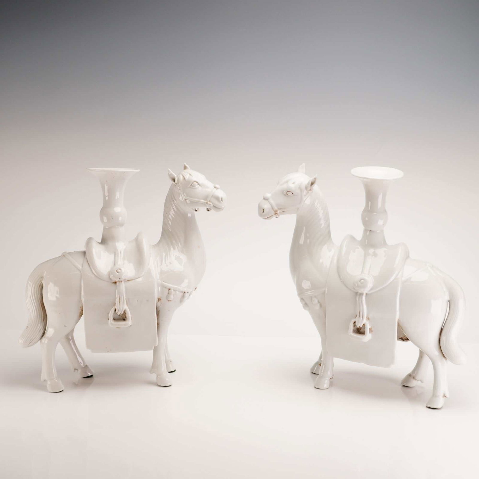 Pair of White Glazed Ceramic Figural Horse Candle Holders: Each horse is modeled in a standing position with the head turned to the side, featuring detailed molded trappings including bridles, tasseled breastplates, and saddles. A trumpet-shaped vessel with