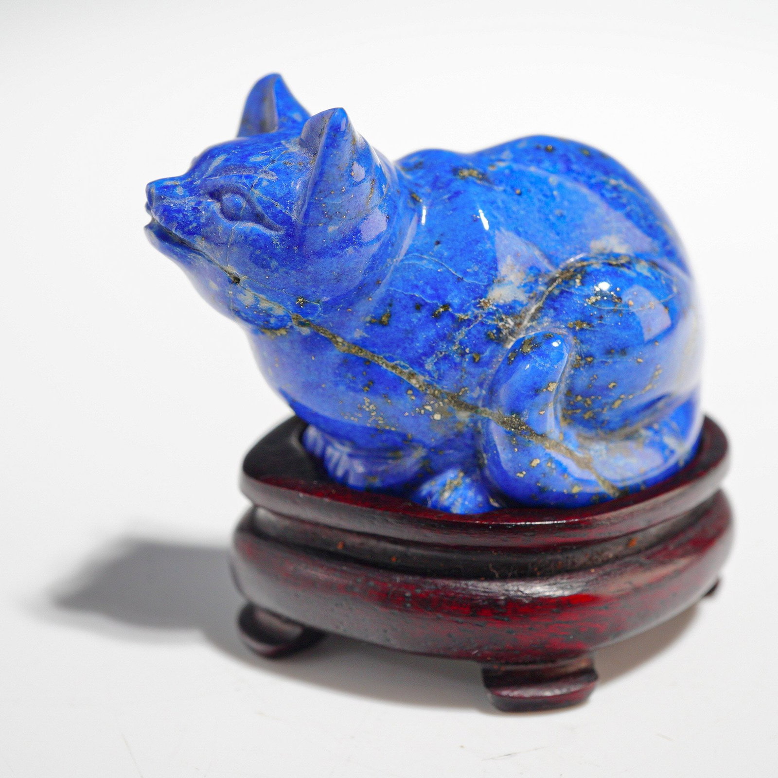A Lapis Lazuli Carving of a Crouching Cat with Fitted Hardwood Stand: This lot comprises two components: a miniature figure of a crouching cat carved from Lapis Lazuli, and its accompanying fitted hardwood stand. The cat figure is executed in a naturalistic style, utili