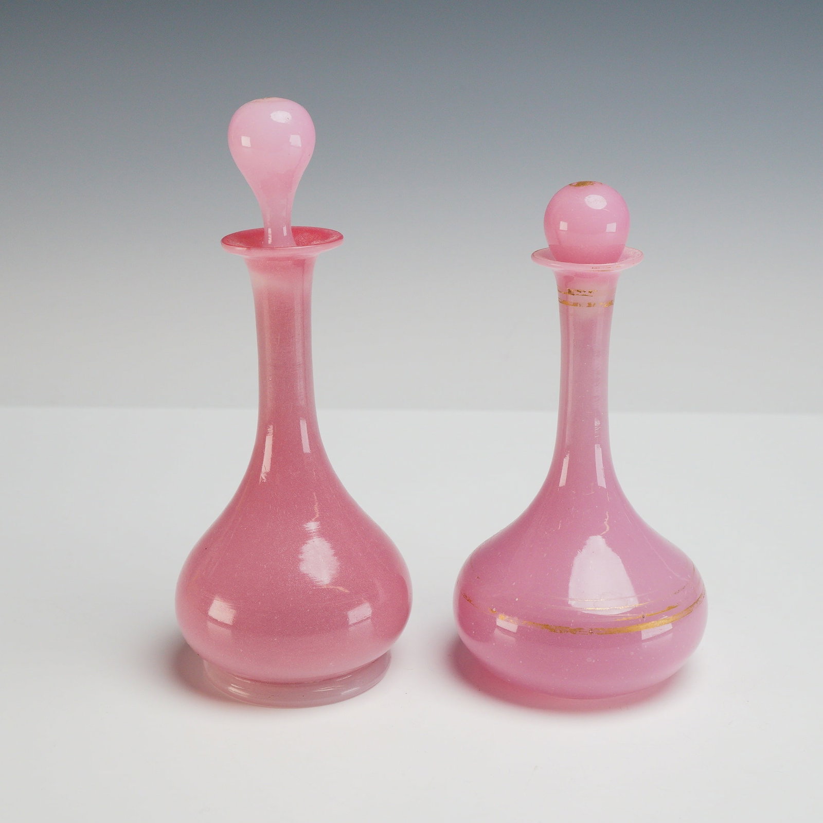2pc Hand-Blown Pink Opaline Glass Scent Bottles with Stoppers: This lot consists of two hand-blown pink glass bottles featuring an opaline or opalescent quality. Each bottle is designed with a bulbous body, a long slender neck, and a flared rim. The taller