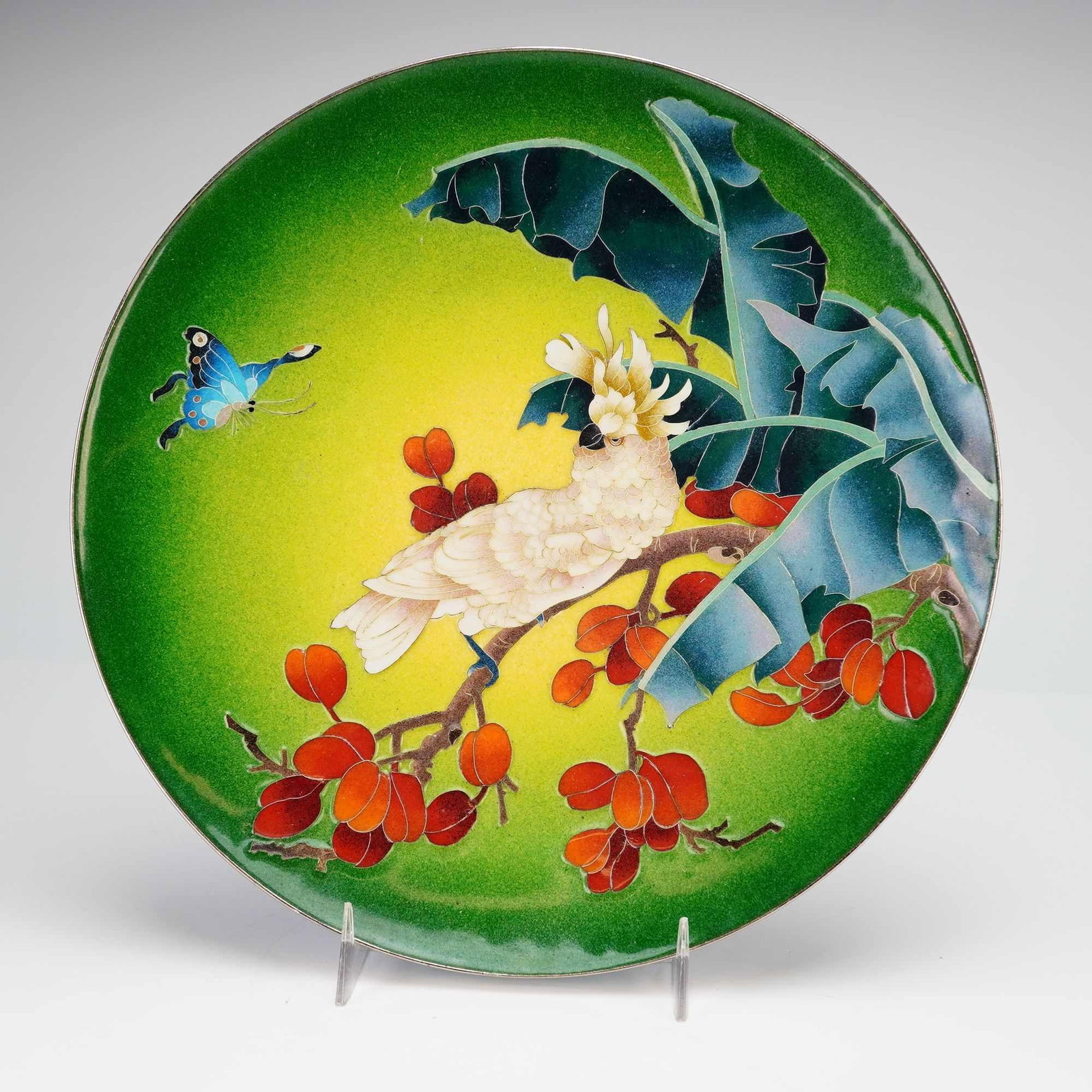 A Contemporary Enamel-on-Metal Decorative Charger with Cockatoo, dated 1984: This contemporary enamel-on-metal decorative charger features a vibrant tropical scene across its face. The composition depicts a white cockatoo with a yellow crest perched on a branch laden with red