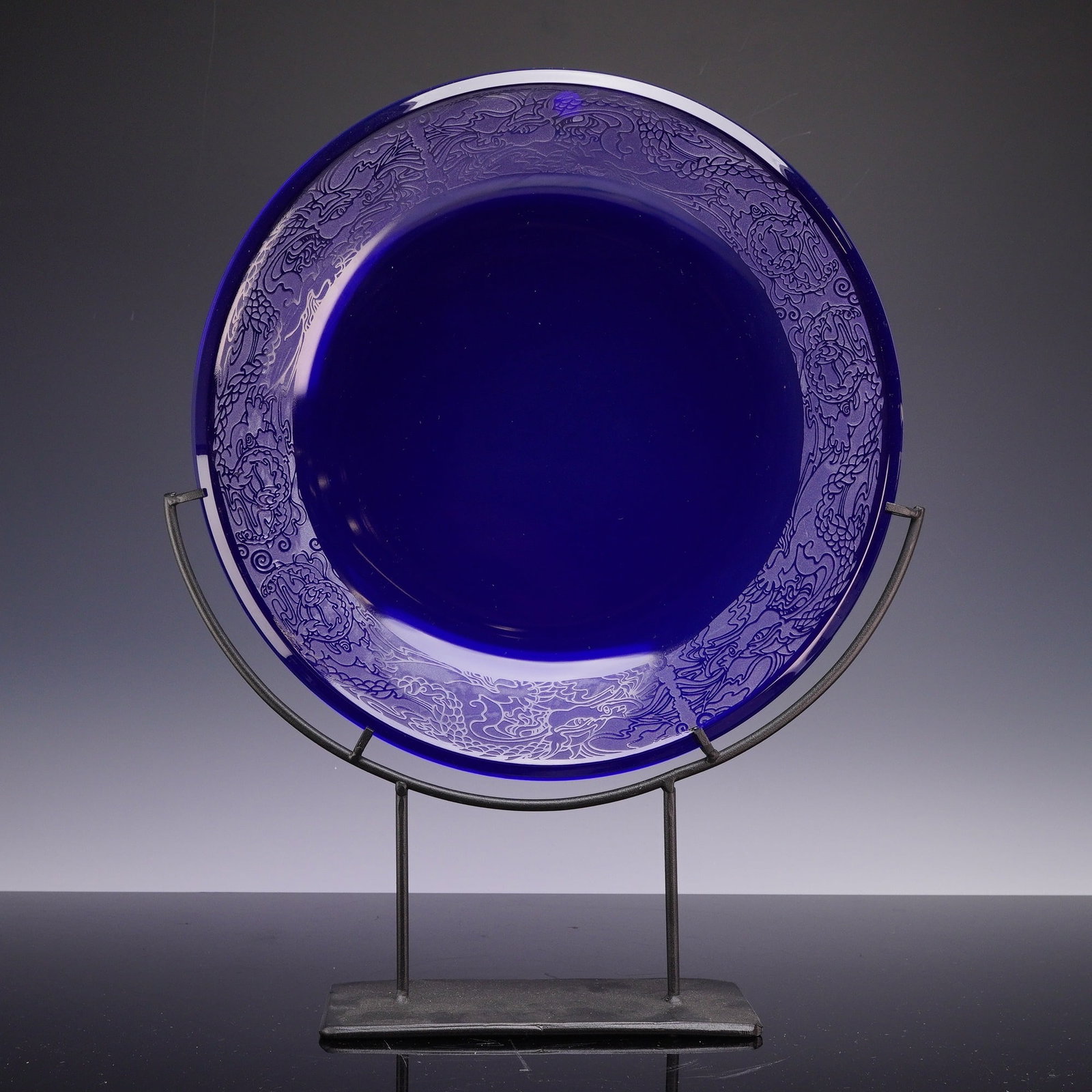 Signed Cobalt Blue Glass Charger and Stand: A decorative cobalt blue glass charger signed by the artist and dated 2010. The piece features a wide rim embellished with a continuous, sandblasted dragon motif. The intricately detailed design