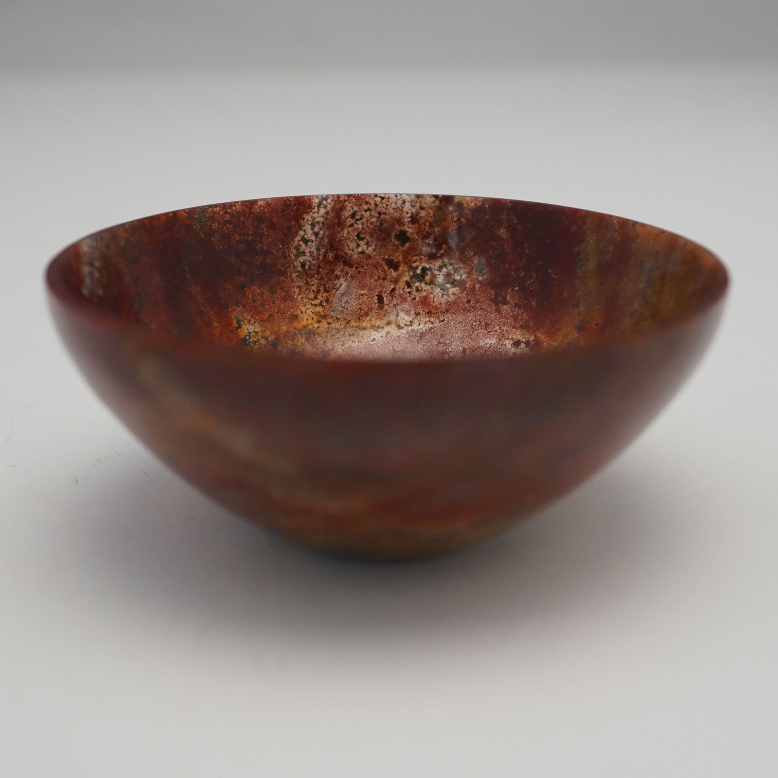 A Polished Jasper Bowl: A small, polished jasper stone bowl, characterized by its naturalistic, variegated material that displays swirling patterns of deep reddish-brown, russet, ochre, and mossy green hues. Its form is a