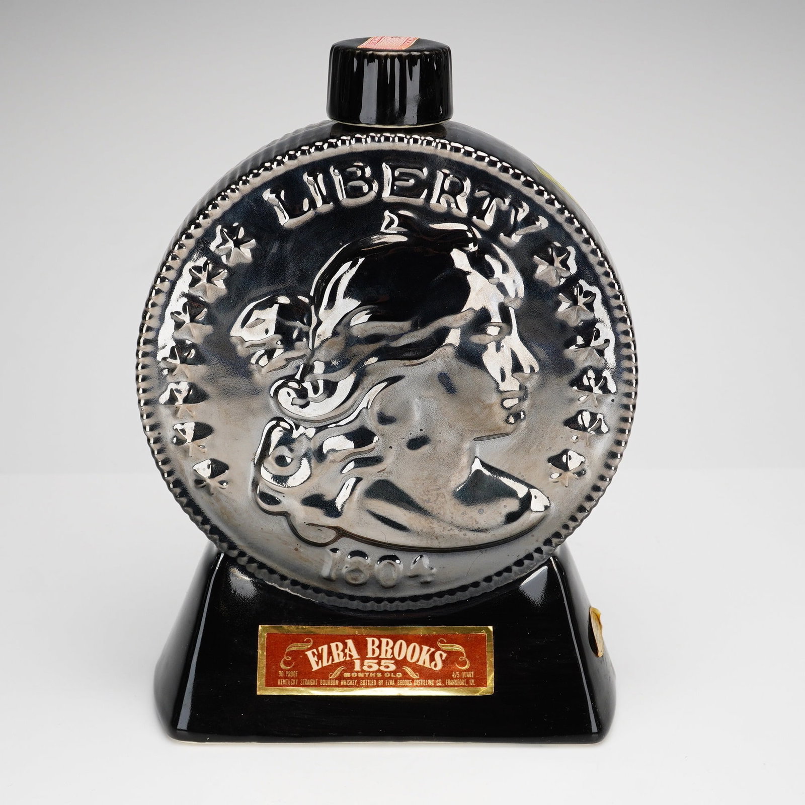 1969 Ezra Brooks Heritage China 1804 Silver Dollar Whiskey Decanter: This ceramic whiskey decanter was produced for the Ezra Brooks Distilling Co. in 1969 as part of their Heritage China series. The vessel is designed in the form of an oversized 1804 Draped Bust Silver