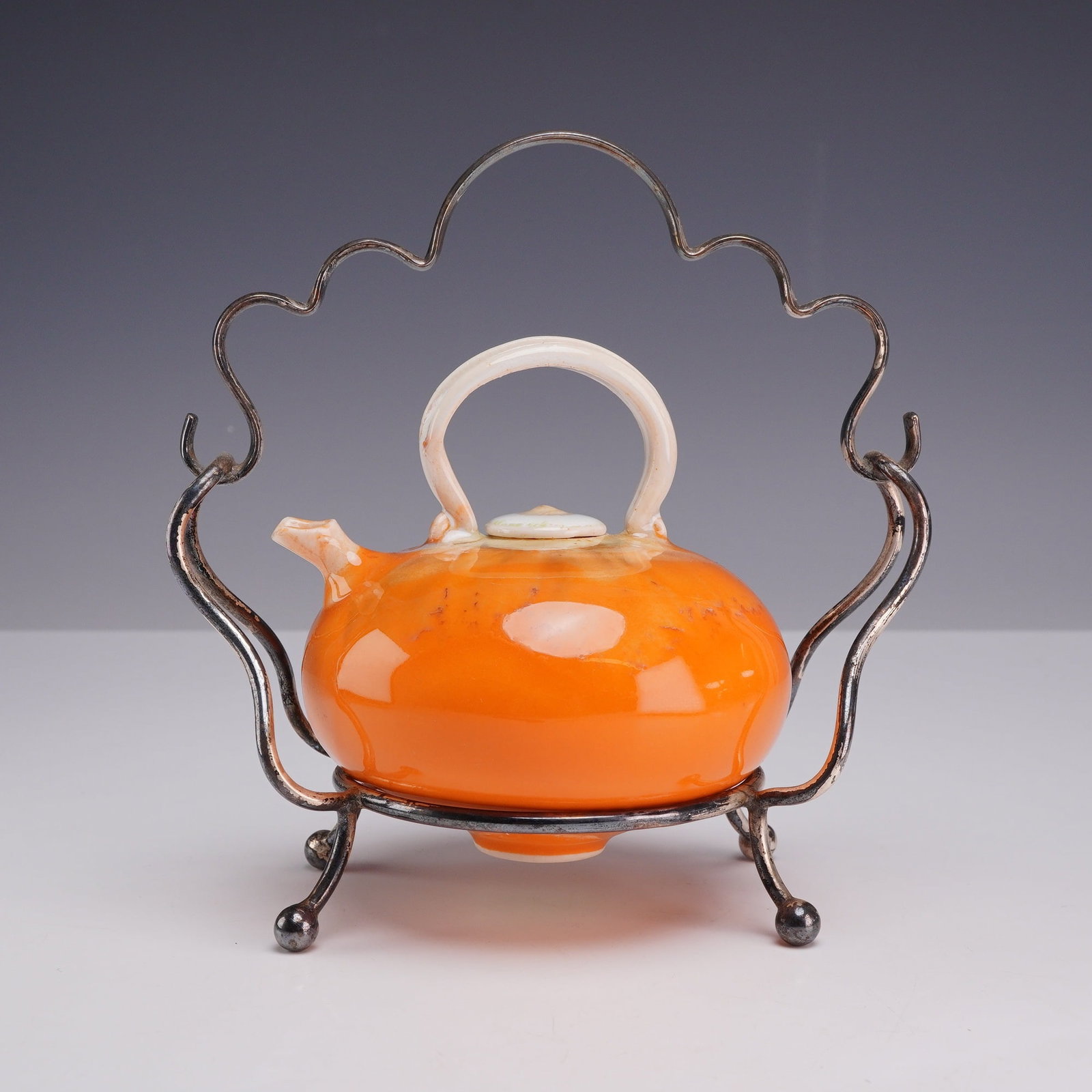 Fong Choo Orange Glazed Ceramic Teapot on Silver-Plated Stand: This ceramic teapot is a product by Fong Choo (Singaporean/American, 20th/21st century). It features a bulbous, squat body finished in a vibrant, high-gloss orange glaze with cream-toned variations co